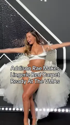 #AddisonRae had a dazzling white swan moment at the 2024 #MTV #VMAs. The white satin bra and panty set lined with scalloped trim was from Miss Claire Sullivan but thanks to some tulle and feathers, the singer took the look from the bedroom to the stage: she added a voluminous tulle bustle that fanned out behind her like a swan and feathers that emerged from her bra and bustle.
 Rae kept accessories simple, adding only a pair of white peep-toe satin pumps, a white hair barrette, and a belly-button piercing to match.