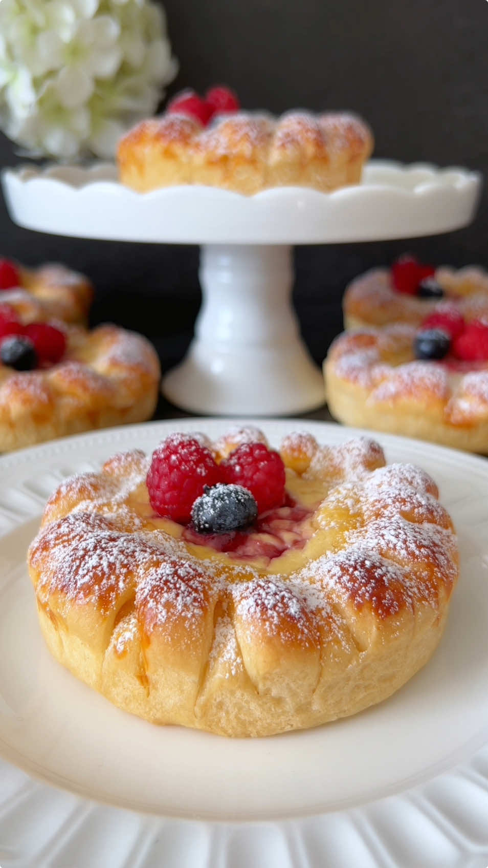 Click here for the Recipe 🫐🤍 Quark Wreaths 🫐 These little delights are not only visually impressive but also delicious with their airy texture and sweet fruity note. Follow each step and enjoy baking! 😍 📝 Ingredients For the yeast dough: •	250 g all-purpose flour (sifted) (2 cups) •	30 g sugar (2 ½ tablespoons) •	1 pinch of salt •	1 tsp dry yeast (0.1 oz) •	120 ml lukewarm milk (½ cup) •	50 g soft butter (3 ½ tablespoons) •	1 egg (M) For the quark filling: •	200 g quark or unsalted cream cheese (¾ cup) •	30 g sugar (2 ½ tablespoons) •	1 egg (M) •	1 tsp vanilla extract •	1 tbsp cornstarch (0.5 oz) For decorating and brushing: •	Jam of choice (e.g., raspberry) •	1 egg yolk, beaten (for brushing) •	Fresh berries •	Powdered sugar for dusting 🤍 Instructions 1.	Sift the flour into a bowl and make a well in the center. Add the yeast, 1 tsp sugar, and 2–3 tbsp lukewarm milk into the well. Cover and let sit for 5–10 minutes until foamy. Then add the remaining milk, sugar, salt, butter, and egg. Knead into a smooth dough. Cover and let rise in a warm place for about 1 hour until doubled in size. 2.	Divide the dough into seven equal balls (about 75 g each). Roll each ball into a circle (about 15 cm). Lightly press a 10 cm baking ring into the center to mark. 3.	Make several small cuts around the mark. Fold each cut strip over in a crisscross pattern (alternating left and right) to create a braided look. 4.	Place the shaped circles into greased 10 cm baking dishes. Cover and let rise for another 30 minutes. Press the cuts down gently with your fingers. 5.	Brush the braided edges with the beaten egg yolk. Add the quark filling to the center of each wreath. 6.	Add three small dots of jam on top of the quark filling and gently swirl with a toothpick to create a marbled effect. 7.	Bake in a preheated oven at 180 °C for about 25 minutes until golden brown. 8.	After baking, cover the warm wreaths with a clean towel and let cool. Dust with powdered sugar and garnish with fresh berries before serving. The quark wreaths are a delicate treat for the afternoon or an elegant addition to any festive table. Enjoy! 🥰