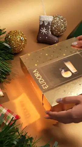 POV: o unboxing de Natal mais lindo, brilhante e cheiroso está passando pelo seu feed! ✨ #Unboxing #Natal #Presentes #GrupoBoticario #PraGeralVer: Em um fundo dourado com decorações natalinas, um kit Eudora Velvet Sensual está sendo aberto. A caixa é dourada e brilhante. No vídeo, mostrando apenas as mãos de uma pessoa, ela passa as mãos pela caixa antes de abri-la; em seguida, ela abre a caixa, pega o frasco da fragrância Eudora Velvet Sensual, dá batidinhas com os dedos, abre a tampa, borrifa no punho e guarda-o na caixa. Depois, pega a embalagem da Loção Hidratante Velvet Sensual, dá batidinhas, aplica uma pequena quantidade na mão e guarda-o na caixa. O vídeo termina com a caixa sendo fechada e retirada de cena.