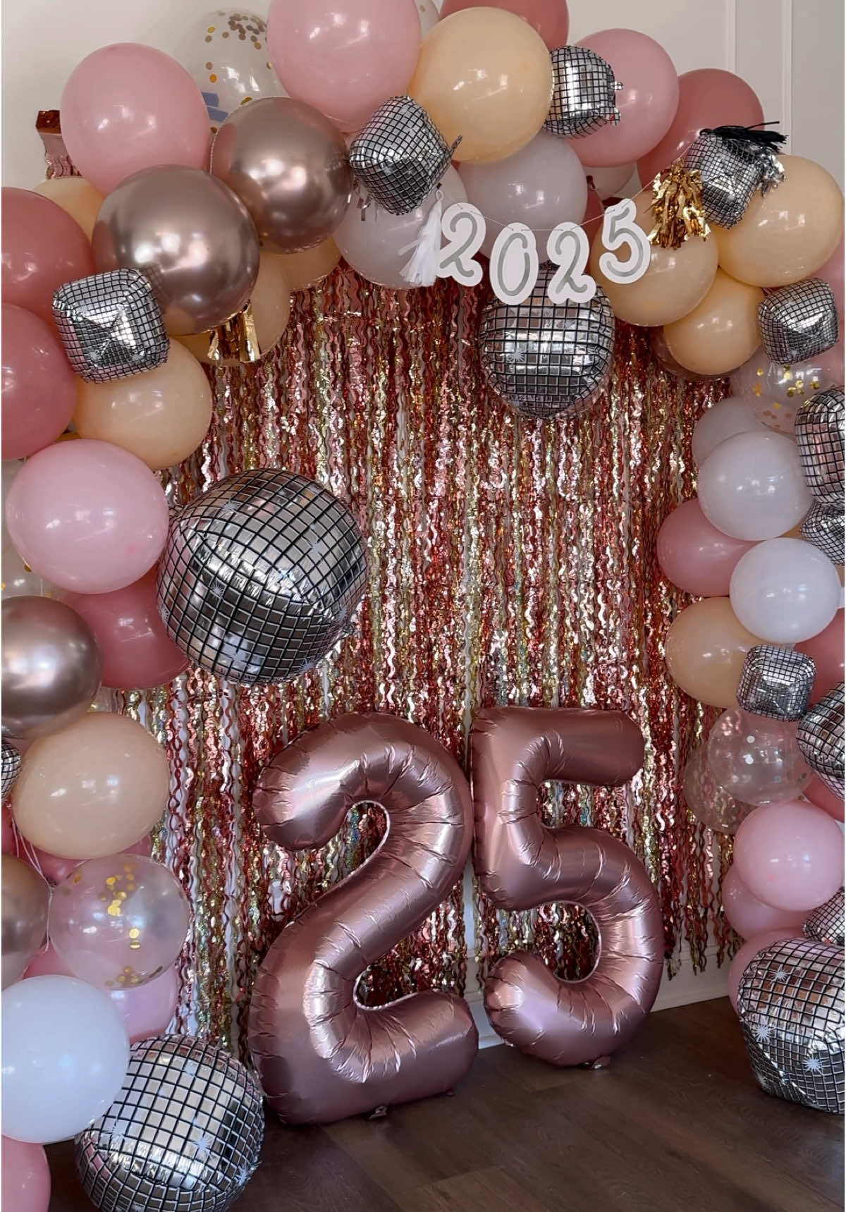 DIY New Year’s Eve party backdrop!!  This was sooo simple to make and is perfect for a New Year’s party! I think in all it took us about 2 hours to make this! @kolterwinton It makes such a cute backdrop for photos with friends and family!! I started with shimmering streamers in the back. I layered gold and rose gold. I wanted to do some brighter colors this year! Then I blew up a bunch of balloons and then tied them together in groups of two and three! Once I tied them together I used a string to intertwine them all! I looped the string around each section of the balloons until it created the full balloon arch! The string is by far the easiest method to hand the balloons! I’ve tried using the balloon kits in the past and they always come out of the balloon holders . Anyways. Once I created the balloon arch with the string , I hung it around the streamers. I used painters tape to tape the string to the wall to hold up the balloons.. then I added some large oversized 25 balloons and 2025 banner across  the front !  And that’s it!! We are Ready for new years!! #newyearseve #diymom #momdiy #momlife #craftymom #newyearseveparty #nye #partyideas #momhack #nye2025 #newyears #funmom #newyearsparty 