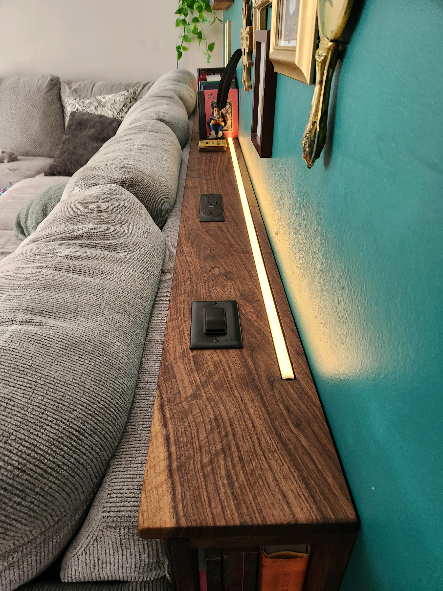 Black walnut sofa shelf with integrated lighting, outlets, wireless charging, and bookshelf I made. This fits perfect with their lounging sofa and artwork on the wall! #custom #custommade #customfurniture #handmade #handcrafted #handcraftedfurniture #woodworking #wooddesign #woodfurniture #interiordesign #interiordesignideas #modern #modernfurniture #midcenturymodern #midcenturymodernfurniture #midcentury #midcenturystyle #midcenturyfurniture #midcenturydesign #fyp #walnut #sofa #couch #bookcase #led #ledlights 