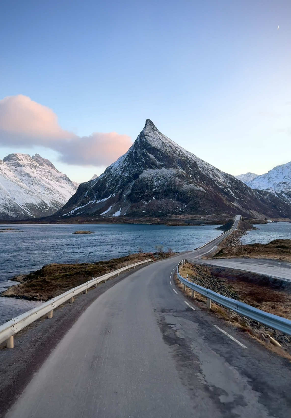 One of the most beautiful roads in the Lofotens in Norway. I had the privilege to rent a camper van for 5 nights and hike some snowy peaks during the days and see the Aurora borealis in the nights. Driving during sunset here from Fredvang with a view of Volandstind mountain. The pink clouds and the moon in the upper right makes the scene so magical for me. #travel #winter #norway #roadtrip #camper #vanlife #fyp #viral #nature #foryoupage #trending #trendingvideo #europe #sunset #mountain 