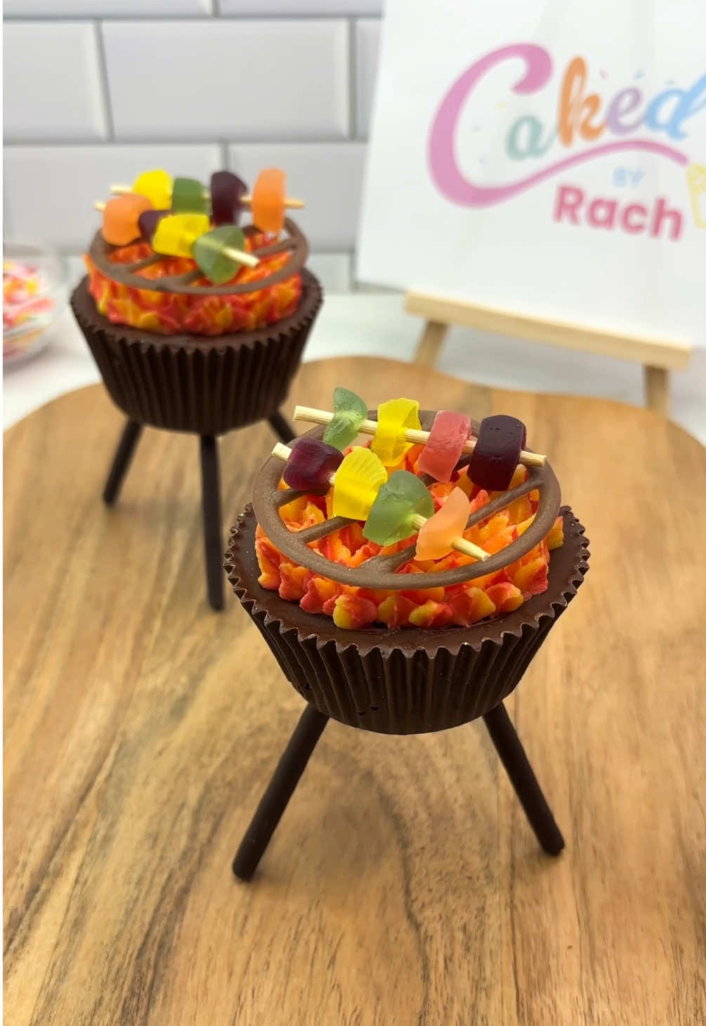 BBQ cupcakes 🔥🧁 You can absolutely eat the whole lot 🤪😵🫶🏻 Taking my edible wrappers to the next level 👀😜 🍫I used melted chocolate to coat the sides of my cupcake liners. I pushed a cupcake into the melted chocolate before it set.  🧁 I attached three pocky sticks for the legs. 🔥 For the flames I used multicoloured buttercream and piping tip 362. 🫶🏻 The grill is melted chocolate that I piped onto parchment paper first.  🍡 The kebabs are chopped up haribo on toothpicks.   Hands up if you remember the first time I made this design? 🙌🏻🤔🫢 #cupcake #cakedecorating #bbq #minifood #cutefood #cakedbyrach #cupcakedesign #grillcupcakes #cupcakesforanyoccasion   