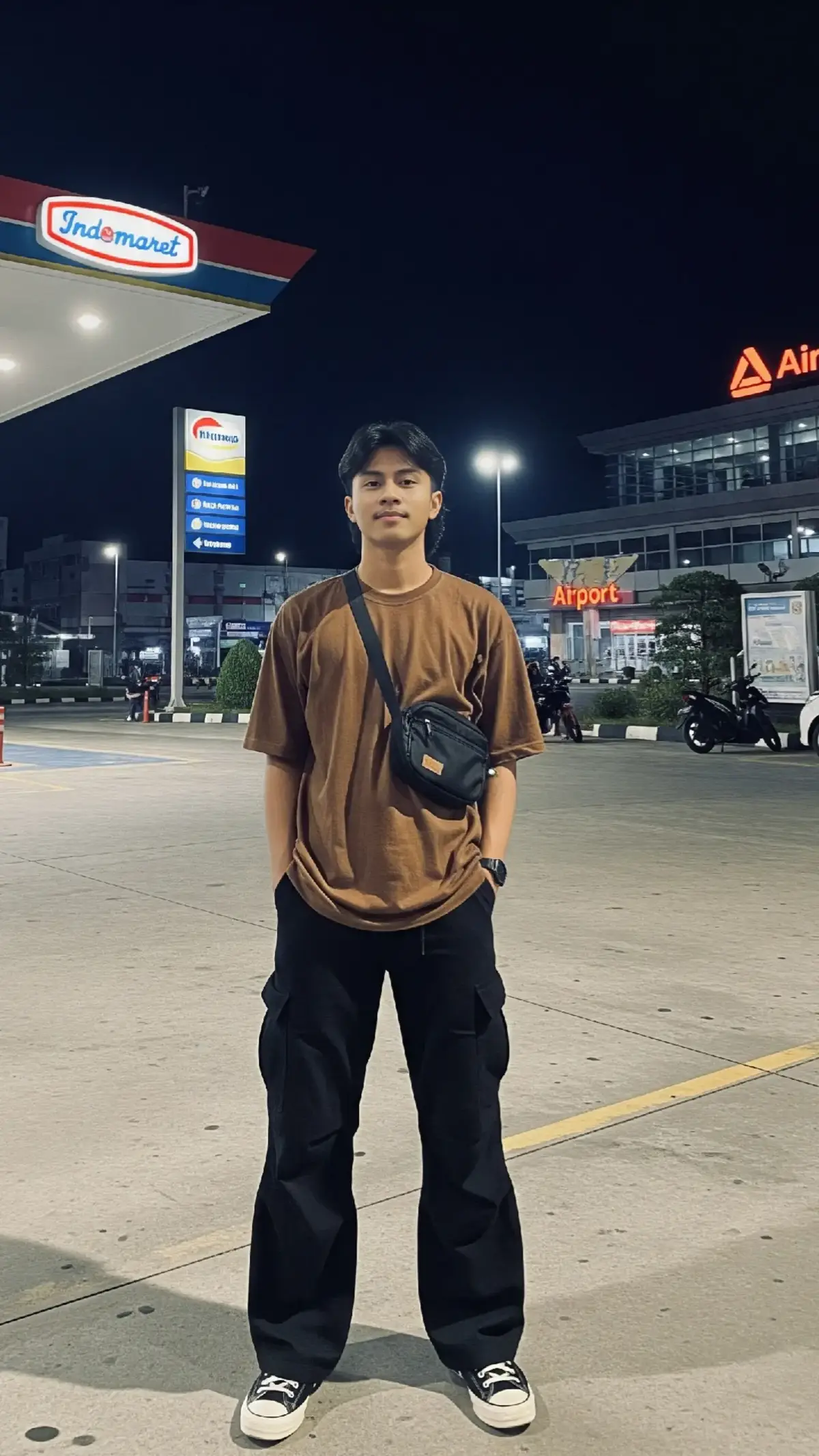 prompt: buatkan foto malam luar ruangan dengan jepretan penuh memperlihatkan seorang pria muda Indonesia umur 18-an berkulit terang dengan rambut hitam gaya model mullet, mengenakan kaos longgar kebesaran warna coklat desain keren, celana cargo longgar kebesaran warna hitam dan sepatu Converse all Star menggendong tas selempang kecil warna hitam ia berdiri tegak dengan kedua tangan ke saku celana tersenyum tipis dan menatap langsung ke kamera ia tampak berada di tempat parkir atau halaman depan pom bensin di belakangnya ada sebuah toko Indomaret yang terang bandara di sebelah kanan dengan berbagai tanda logo terlihat di sebelah kirinya sebuah mobil putih terparkir di sebelah kanannya sebuah sepeda motor hitam terparkir di latar belakang ada bangunan lain dan langit gelap pencahayaan keseluruhan buatan dan terang di sekitar toko menerangi pria dan latar depan resolusi 3:4 #fyp #ai #fypシ゚viral #lewatberanda 