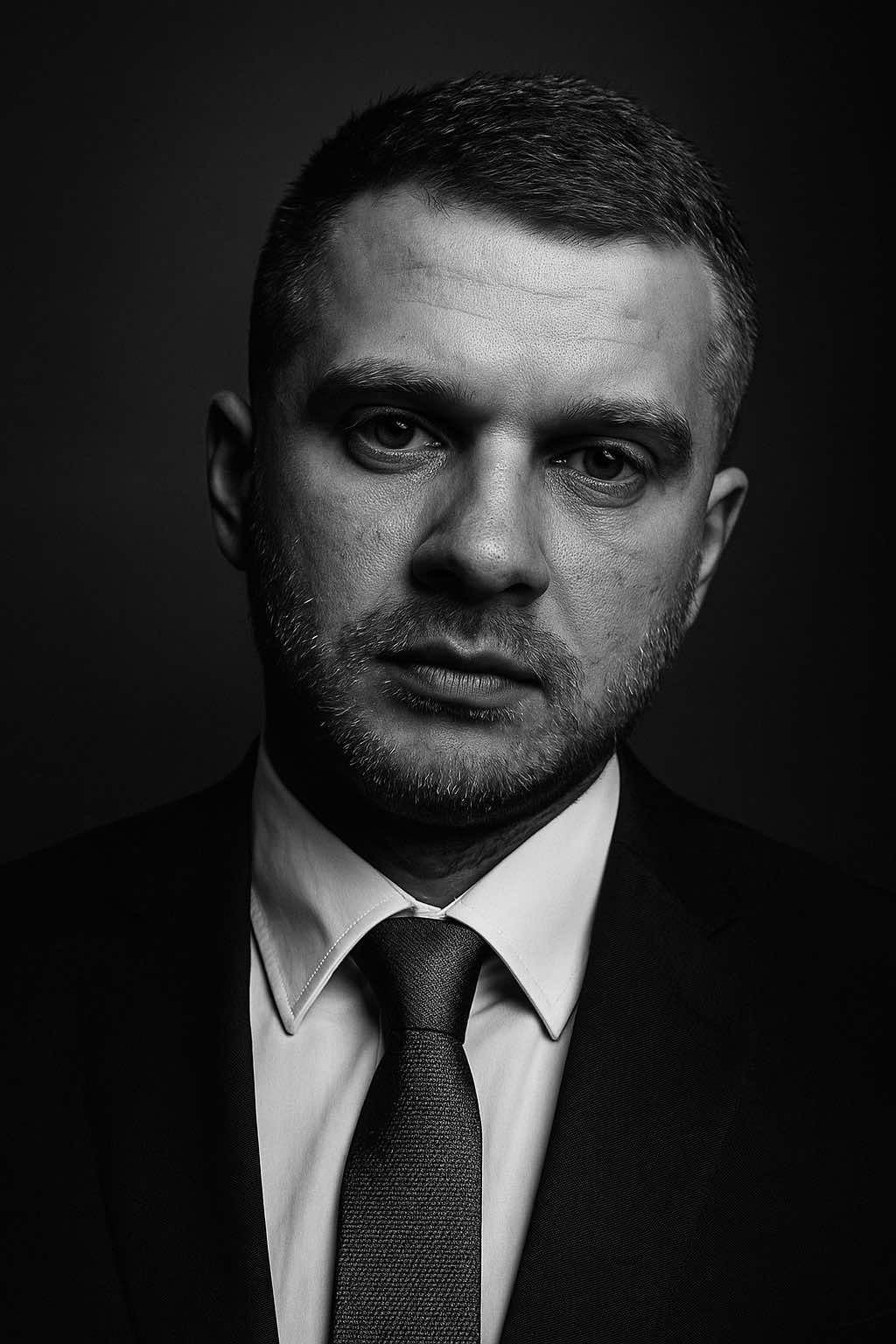 B&W portrait ChatGPT photo idea. P.5 Your thoughts?  Create a high-end black and white portrait using my selfie. The face must remain exactly as in on my original selfie — no editing, no retouching, no smoothing. I’m wearing the business suits. Preserve every facial detail and texture for a raw, authentic look. The lighting should be dramatic and studio-quality, using strong contrast and shadows to sculpt the features. Use a clean, blurred studio-style background with soft gradients or subtle texture to enhance focus on the subject. Frame the portrait vertically (9:16 format), centered composition, with ultra-realistic skin texture and depth. No digital makeup or alterations. The final result should look timeless, editorial, and emotionally powerful — suitable for a gallery or fashion magazine.