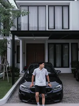 PROMPT:gambar berasio 9:16 dan gambar diambil menggunakan kamera/drone. menampilkan seorang pria berusia 17 tahun,berbadan ideal tinggi 170 bb 55 dengan badan yang bersandar menghadap kedepan pada sebuah mobil bmw seri m4 hitam yang terparkir di carport salah satu unit rumah mewah modern dua lantai bergaya minimalis.Rumah tersebut memiliki atap datar,jendela besar untuk pencahayaan alami,jalan paving block,dan area hijau yang tertata.pria itu berkulit sawo matang terang disebelahnya,menggunakan kaos polo pendek putih,mengenakan celana pendek hitam,dan sepatu putih,