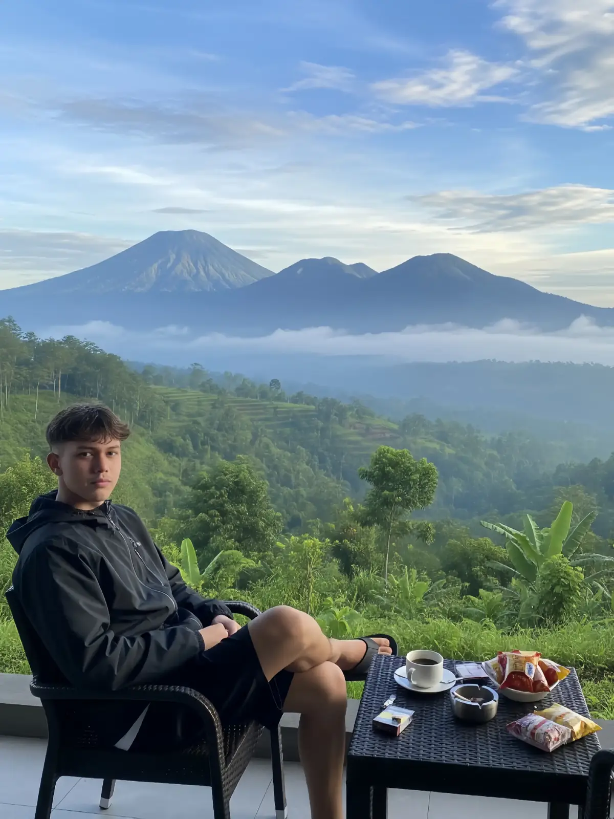 prompt. : Very beautiful and realistic tropical natural scenery, cloudy peaks, Indonesia, with clouds covering the mountains in the morning creating a sunrise atmosphere, the mountains covered with clouds look beautiful, white clouds hanging in the bright blue sky, and trees full of mist. In the foreground, a 25-year-old white Indonesian man, about 175 cm tall, fair skinned, black hair with a neat undercut, facing the front camera with a slight smile, sitting on a chair in a villa, complete with a table containing, ashtray, cigarettes, snacks, and a cup of hot coffee, with a beautiful mountain background. He is wearing a thick black parasite jacket, oversized black outdoor shorts, and black flip-flops. Natural lighting from the morning sunrise, soft shadows on the body and vegetation, a slightly above angle (high-angle shot) to capture the beauty of the landscape and the man's pose dramatically and blend in with nature. The aspect ratio is 3:4 #fyp #ai #trend #gemini #geminiai #trending #foryoupage 