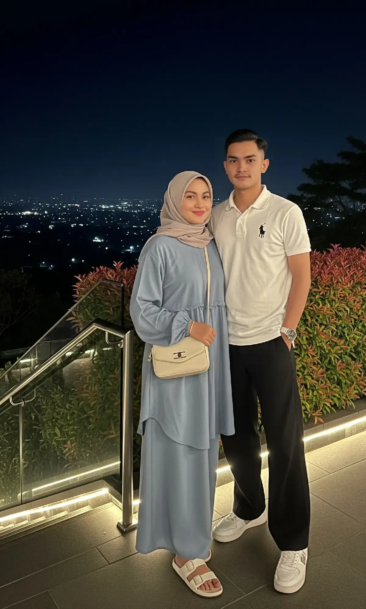 Promt Gemini :A young Sundanese couple are outdoors at night. Main Subject: The Sundanese woman She is wearing a dusty blue gamis (dress) with loose sleeves, a matching hijab in a simple and elegant style, carrying a small off-white sling bag with metallic accents on the front, and wearing white sandals. She poses with a sweet smile, looking at the camera, her body slightly turned to the side. The Sundanese man He is wearing a white polo shirt with a small black horse logo on the chest, black wide-leg pants, a white watch, and chunky white sneakers. Background: The location is outdoors at night. In the distance, a cityscape with twinkling lights can be seen. Beside them is a glass railing and iron railing on the side of the stairs, illuminated from below with warm lights. Behind the couple is a green hedge with reddish leaf tips and tall trees silhouetted against the night sky. @Ariel Tatum  #gemini #chatgpt #faceswap #ai #klingai #googlegemini #promt #arieltatum #fypシ゚ #aiphotography #trendddddddddddddddd #trendingggggggggggggggggggggggggggggggg #fyppppppppppppppppppppppp #viralllllllllllllllllllllllllllllllllll #xycbaa #fouryou #xycbaaaaaaaaaaaaaaaaaaaaaaaaaaaa #xyczba #trending #aiphoto #trending?song🔥 
