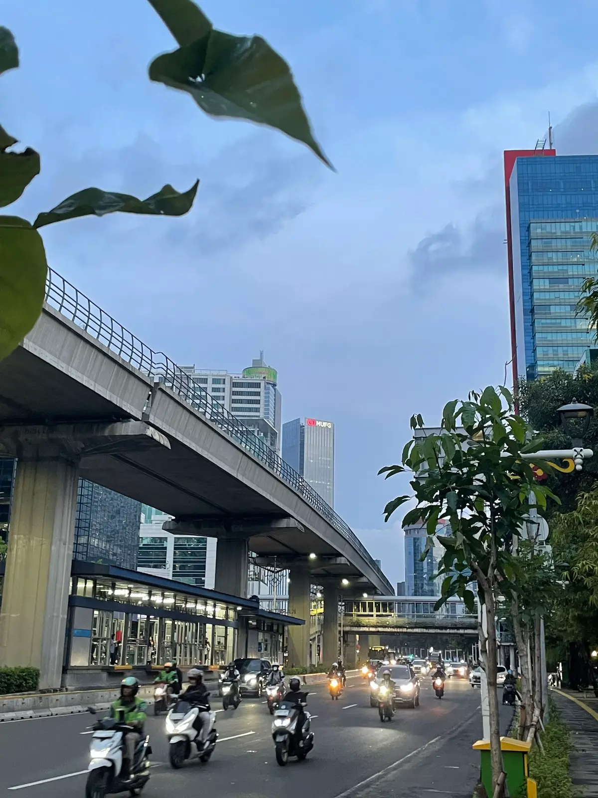 Foto-foto ini diambil pada waktu sore hari, saat matahari mulai turun dan langit Jakarta berubah warna. Lokasi pengambilan gambar berada di kawasan Kuningan, tepatnya di sekitar Setiabudi, Jakarta Selatan — sebuah kawasan bisnis yang selalu hidup, bahkan ketika hari mulai gelap. Dengan menggunakan kamera handphone iPhone 12, saya mencoba menangkap suasana kota dari berbagai sudut pandang, memanfaatkan pencahayaan alami senja dan karakter arsitektur kota. Dalam gambar pertama yang dijadikan sampul, terlihat suasana jalan layang yang membentang di antara gedung-gedung tinggi. Di sisi kiri bawah, ada stasiun LRT Setiabudi Astra yang tampak rapi dengan pencahayaan hangat dari lampu-lampu jalan. Langit pada gambar ini menunjukkan transisi antara biru siang menuju jingga senja, memberi kesan tenang sekaligus dinamis — sangat khas Jakarta di akhir hari kerja. Foto kedua menyorot sisi lain dari gedung pencakar langit dengan sudut pandang dari bawah ke atas. Pohon-pohon palem di latar depan memberi kontras yang unik terhadap dinding kaca gedung, menciptakan perpaduan antara unsur alam dan kemajuan teknologi kota. Refleksi cahaya matahari yang tersisa tampak halus pada permukaan kaca, menciptakan efek visual yang lembut namun tegas. Gambar ketiga menampilkan jembatan penghubung yang melintas di antara dua area, dengan latar belakang langit biru cerah yang mulai memudar. Komposisi ini terasa seimbang — menghadirkan struktur horizontal jembatan di tengah bingkai dan bayangan pepohonan di bagian atas. Suasana ini menggambarkan perpaduan mobilitas dan kehijauan kota yang jarang kita sadari saat terburu-buru menjalani hari. Foto keempat menangkap pemandangan luas ke arah kawasan perkantoran Setiabudi. Bangunan-bangunan tinggi berdiri megah di kedua sisi jalan, sementara langit tampak masih cukup terang walaupun hari mulai sore. Lampu-lampu mulai menyala, memberikan kesan kontras yang indah antara cahaya buatan dan cahaya alami yang mulai redup. Warna merah di puncak salah satu gedung menambah aksen visual yang menarik, menunjukkan betapa kaya warna sore hari di Jakarta bisa tercipta hanya dengan kamera ponsel. Keempat foto ini saya ambil hanya dengan menggunakan kamera iPhone 12 — perangkat yang mungkin sudah akrab digunakan oleh banyak orang, namun tetap mampu menghasilkan detail, pencahayaan, dan kontras yang mengagumkan jika digunakan pada waktu yang tepat. Sore hari menjadi pilihan terbaik untuk mengambil gambar karena pencahayaannya yang lembut, tidak terlalu keras seperti siang hari, dan mampu menciptakan gradasi warna langit yang dramatis. Sudut-sudut pengambilan gambar saya sesuaikan untuk menangkap elemen-elemen vertikal dan horizontal yang kuat di kawasan ini. Dengan mengambil gambar dari sudut bawah (low angle), saya ingin menampilkan megahnya bangunan tinggi yang sering kali luput dari pandangan kita sehari-hari. Sedangkan sudut lurus atau eye-level saya gunakan untuk menangkap komposisi jalan dan jembatan, sehingga menampilkan kedalaman dan keseimbangan visual dari suasana kota. Setiabudi dan Kuningan adalah kawasan yang terus berkembang, penuh dengan aktivitas bisnis, namun tetap memiliki spot-spot estetis yang bisa dinikmati, bahkan hanya dengan berjalan kaki dan membawa kamera ponsel. Area ini memiliki banyak elemen visual menarik — dari jalan layang, pohon palem, gedung kaca, hingga jembatan penyambung — yang semuanya bisa menjadi objek fotografi urban yang menarik. Foto-foto ini bukan hanya dokumentasi visual, tapi juga refleksi kecil tentang bagaimana kita memandang kota. Jakarta, meskipun padat dan sibuk, tetap menyimpan keindahan di sela-sela keramaiannya. Dengan sedikit waktu luang dan perhatian pada detail sekitar, kita bisa melihat wajah lain dari kota ini yang tidak kalah menawan.