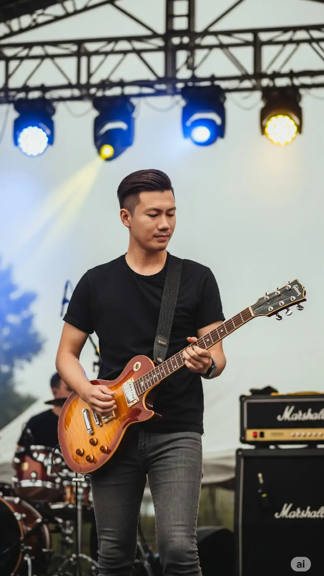 Potret realistis seorang pria muda Asia memainkan gitar elektrik dalam suasana konser outdoor di siang hari yang mendung. Ia mengenakan kaus hitam polos dan celana jeans slim fit berwarna abu gelap, dengan gitar sunburst 6 senar yang digantung dengan strap hitam, memiliki gaya rambut pendek rapi yang disisir ke samping. Ekspresinya ke arah samping kanan, tenang dan fokus ke arah penonton, menunjukkan kesan profesional dan percaya diri. Latar belakang menunjukkan panggung konser outdoor dengan lampu sorot biru dan kuning menyala di belakangnya, serta struktur rigging logam panggung terlihat jelas. Cahaya ambient alami dari langit mendung menciptakan tone warna soft dan sedikit moody. Di belakang, terlihat samar-samar seorang drummer dan perangkat drum set serta amplifier Marshall besar berwarna hitam. Suasana keseluruhan menunjukkan momen live music performance yang energik namun tetap fokus, dengan detail instrumen, pencahayaan panggung, dan nuansa realistis yang tajam. Gaya foto menggunakan lensa aperture besar (depth of field sempit), bokeh latar belakang lembut, fokus tajam pada subjek utama, kualitas ultra-realistik 8K, pencahayaan natural dan warna tone hangat netral. Resolusi 9:16 #ai #chatgpt #fotoai #gemini #mbw 