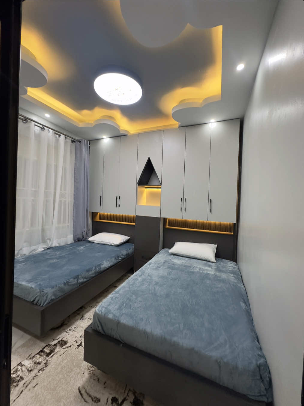 This Twin Bedroom Setup Designed with both function and fantasy in mind, this setup makes every square inch count. Two identical beds sit perfectly parallel, separated by a stunning custom wardrobe that anchors the room in symmetry and structure. We didn't just build storage, we sculpted it. The central wardrobe features full-height cabinetry, giving plenty of room to tuck away clothes, toys and clutter. A triangle-shaped display niche with soft ambient lighting adds a geometric twist that feels both modern and magical. From the open shelf to the sleek handle design, every element was thought through with purpose. Look up and you'll find the ceiling steals the show, With its playful cloud-shaped gypsum curves, LED cove lighting, and a custom Paw Patrol light feature, it's a ceiling that tells a story even before bedtime begins. We went for rich textures — a neutral-toned abstract rug for warmth underfoot, and a palette of soft greys and calming blues that grow with the kids, without ever growing dull. It's smart. It's stunning. And it's saving space in the most stylish way possible. If you've got little ones and a not-so-little love for good design, This is your sign to book us. DM to transform your kids' room into something unforgettable. 0751 200 500 0750 200 500 #BossInteriors #KidsRoomGoals #TwinRoomDesign #SpaceSavingInterior #InteriorDesignForKids #FunctionalAndBeautiful #SharedRoomInspo