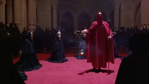 هل فلم Eyes Wide Shut يتكلم عن الماسونية؟  Eyes Wide Shut  هو آخر أفلام المخرج العبقري ستانلي كوبريك، وهو عمل سينمائي يختلط فيه الغموض بالرمزية النفسية والاجتماعية. القصة تتبع الدكتور بيل هارفورد (توم كروز) الذي يعيش حياة هادئة نسبيًا مع زوجته أليس (نيكول كيدمان) حتى تعترف له ذات ليلة بأنها شعرت بانجذاب لرجل غريب. هذا الاعتراف البسيط يهز كيانه ويشعل بداخله فضولًا جامحًا يدفعه للخروج في رحلة ليلية عبر نيويورك، رحلة ستقوده إلى عتبة عالم غامض لم يكن يتخيله. في أثناء هذه الرحلة، يصل بيل إلى قصر ضخم تقام فيه طقوس سرية، يشارك فيها أشخاص من الطبقات العليا، جميعهم يرتدون أقنعة فاخرة ويرقصون وسط أجواء مشحونة بالإغراء والخطر. المشاهد المليئة بالرموز، الطقوس الغامضة، والموسيقى الكنسية جعلت كثيرين يربطون الفيلم بالماسونية أو الجمعيات السرية، لكن الحقيقة أن كوبريك لم يصرح أبدًا بأن العمل يفضح الماسونية أو يتحدث عنها مباشرة. بدل ذلك، يستخدم كوبريك هذه الطقوس كأداة رمزية للتعبير عن عدة أفكار: السلطة والنفوذ: كيف تعيش النخبة في عوالم مغلقة لا يمكن لعامة الناس الوصول إليها. الرغبة البشرية: كيف يمكن أن تقود الغيرة أو الفضول الشخص إلى اتخاذ قرارات خطيرة. الغموض الاجتماعي: أن هناك دائمًا ما هو خفي وراء الواجهة الأنيقة للمجتمع. الفيلم ليس عن الماسونية، بل عن فكرة 