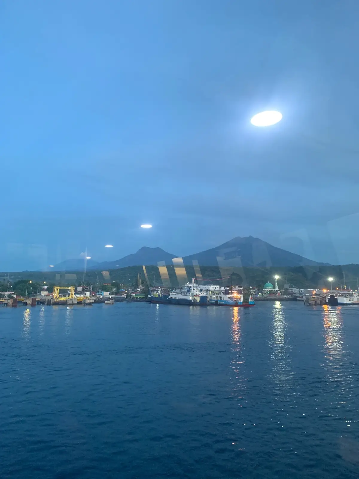 Pelabuhan yang sibuk dengan kendaraan menanti penyeberangan, dikelilingi pemandangan laut biru yang tenang dan pulau hijau nan asri. Suasana tropis yang menyejukkan mata dan hati, anjayyy 🦦