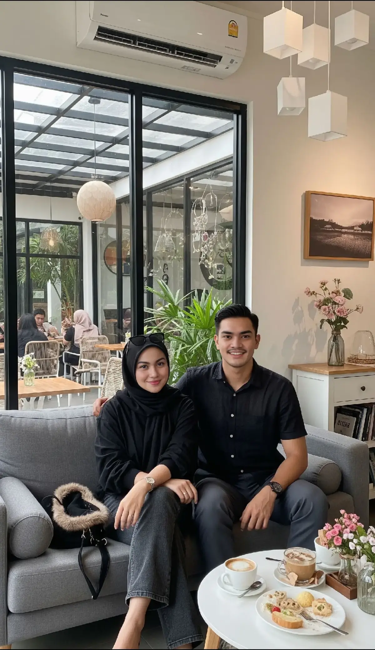 Promt Gemini : Main Subject: A woman wearing a black hijab, wearing a loose black shirt with rolled-up sleeves and faded pastel black trousers. She sits casually on a gray sofa, smiling at the camera, and poses with relaxed hands. She is seen wearing a thin bracelet on her left hand, large sunglasses, and high heels. A padded black bag is placed to the left of the sofa near her. Beside her sits a man wearing a short-sleeved black linen shirt, wide-leg dark gray trousers, and sneakers. His hands are relaxed and his expression is smiling. A metal watch is visible on his left hand and a black bracelet on his right. Interior Setting: An indoor space with large glass walls reveals an outdoor area with cafe tables and chairs, green plants, a glass ceiling with a black frame, and several other people. Inside, there is a gray sofa where they sit, a flower pot, a small white table next to it with food and coffee, a white bookshelf beside it, and a wall decoration consisting of a horizontal grid painting. There are round ornamental plants inside the room. The ceiling of the room is decorated with a modern pendant lamp in the shape of a long white beam. Air conditioning is available. Lighting: Natural sunlight entering through the glass, combined with warm indoor lighting, creates a warm and realistic atmosphere. 3:4 ratio @Ariel Tatum  #gemini #ai #fyp #fomo #jawa 