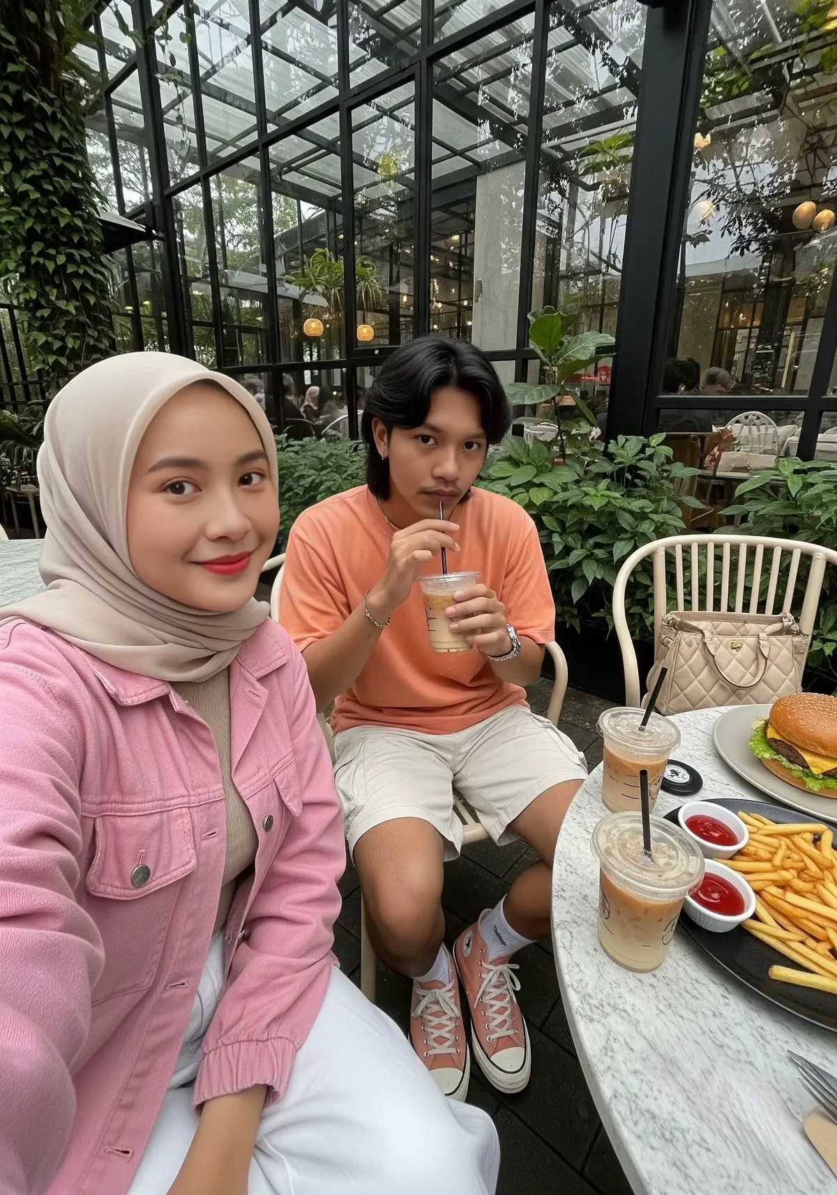 Generated By Gemini . . . . .PROMPT GEMINI : Selfie photo in an outdoor café with lush greenery, modern glass windows, and a cozy aesthetic vibe in the background. A young Indonesian woman wearing a light beige hijab, a pink jacket, and white wide-leg pants is holding the phone slightly above eye level to take the selfie. She smiles warmly toward the camera. Next to her, a very handsome 18-year-old young Indonesian man with a mullet hairstyle (black hair, top part parted down to the forehead) is wearing a dark peach t-shirt, cream cargo shorts, and dark peach Converse high-top sneakers. He poses naturally while drinking iced coffee latte with a straw, glancing coolly at the camera. On the café table visible in the foreground are one cups of iced coffee latte (one in his hand), a plate of french fries with dipping sauce, a large cheeseburger, and a BMW car key. A beige luxury handbag is also partly visible on the chair beside them.Rasio 3:4 #nongkrong #dateideas #couple #gemini #fyppppppppppppppppppppppp 