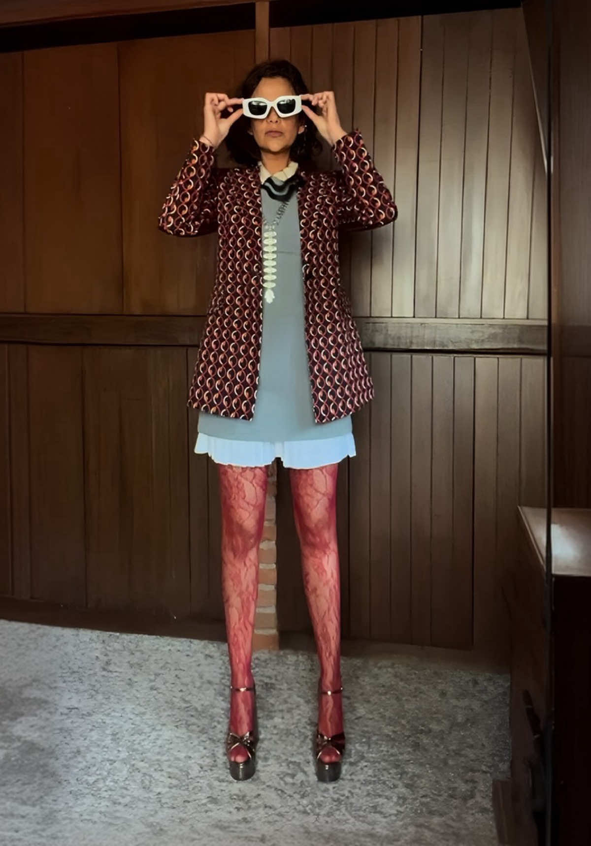 Chaqueta geométrica en tonos vino con patrón retro, combinada con un vestido corto gris de caída ligera y terminado en volantes. Las medias rojas de encaje introducen un contraste inesperado frente a la neutralidad del gris, mientras que las plataformas metálicas en burdeos acentúan la fuerza del conjunto. El collar negro rígido aporta un detalle gráfico y los lentes blancos de armazón grueso subrayan la actitud audaz y contemporánea. Un look que equilibra sobriedad y riesgo en una propuesta con personalidad propia. #fashioninspiration #StreetStyle #StyleWithoutAge #VintageTwist #OOTD