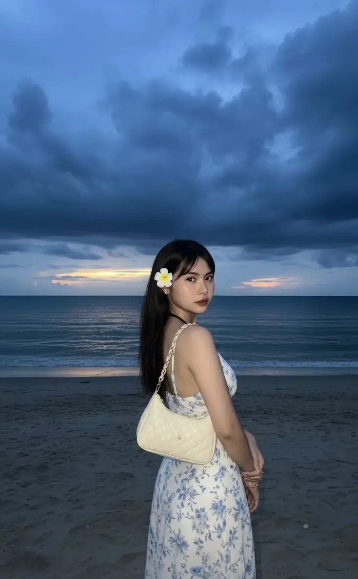 A stunningly realistic sunset beach scene with a dramatic deep blue sky filled with dark clouds. A small amount of light penetrates near the horizon, casting a faint golden reflection on the calm waters. The beach is dimly lit by the afterglow of dusk, creating a serene and somber atmosphere. In the foreground stands a young woman with her back to the camera. She is incredibly beautiful, with flawless, glowing skin. Her face is softly lit by the camera's flash, creating a beautiful contrast against the dark background. She wears her hair down neatly, complemented by a fresh white frangipani flower tucked behind her ear, adding a tropical and feminine touch. Her outfit consists of a white dress with a delicate white floral pattern on a soft blue fabric, evoking a fresh beach look. A quilted ivory shoulder bag with a smooth and soft texture hangs casually on her shoulder. She accessorizes it with a delicate gold bracelet on her wrist and a minimalist ring, while holding both hands in front of her chest. Her relaxed posture elegantly conveys confidence and poise, perfectly blending fashion and nature. The overall image combines highly realistic textures, natural lighting, and atmospheric beach scenery to create a visually captivating and peaceful portrait. 9:16 ratio. Don’t change the face #ai #fyppppppppppppppppppppppp #for #foryoupage❤️❤️ #trending 