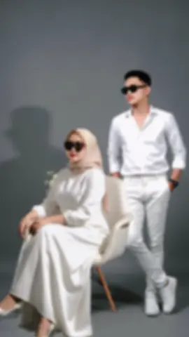 jadikan satu frame foto dua orang ini layaknya seperti foto studio bareng background putih, dengan pakaian wanita muda memakai blezer putih rok panjang putih sepatu heels putih kacamata hitam duduk santai dikursi dan pria muda memakai kemeja putih celana panjang putih sepatu putih kacamata hitam berdiri disamping wanita, dengan pose tubuh yang rileks dan santai elegan namun berwibawa dengan pencahayaan lembut dan minim menghasilkan bayangan tegas dan suasana moodys. estetika sederhana dan modern