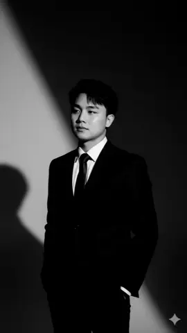 A high-resolution, black-and-white portrait of a young man wearing a sharp black suit and black shirt with a tie. He stands confidently in a minimal studio setting, facing slightly to the side. Dramatic studio lighting casts bold geometric shadows across his face and background, with a strong diagonal beam of light cutting through darkness. His expression is calm, introspective, and slightly distant. The image has a high-fashion, cinematic noir tone, with high contrast and fine detail on the face and suit texture. Soft bokeh background, vertical frame (9:16), 8K resolution.#CapCut 