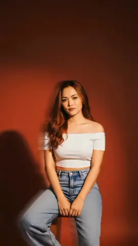 PROMPT:  Create a photo of the above image of a young woman that looking to the camera with solid saturated crimson red background. She was sitting on a wooden stool. Copy the facial features. Make it more realistic. She is wearing a 3 inch white heels. The woman has waist length long wavy hair, that cascade over her shoulder and down her back. She is wearing a white off-shoulder crop top and denim baggy jeans.The lighting casts a soft shadow on the wall behind her. Her expression is serene and peaceful, The mood of the photo is soft and celebratory. #geminiaiprompt #geminiai #fyp #fypシ #trending 