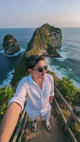 prompt pantai kelingking: Buatkan foto seorang pemuda berswafoto saat mendaki di jalur sempit di tebing berpagar kayu, tanpa mengubah wajah menghadap formasi batuan ikonis berbentuk T-Rex di Pantai Kelingking, Nusa Penida, Bali. Pria mengenakan kemeja putih longgar dan celana pendek putih dengan sandal krem dan kacamata hitam, senyuman manis di bawah sinar matahari keemasan. Laut di latar belakang berwarna biru tua dengan ombak yang menghantam tebing. Vegetasi hijau rimbun mengelilingi jalur. Perspektif swafoto sudut lebar, sudut tinggi, komposisi lengan terentang. Pencahayaan alami yang hangat, bayangan lembut, cahaya keemasan, warna cerah, tekstur realistis, gaya fotografi perjalanan. #GeminiAl #EditFotoAI #AIGeminiTutorial #CaraEditFoto #AIGeneratedContent