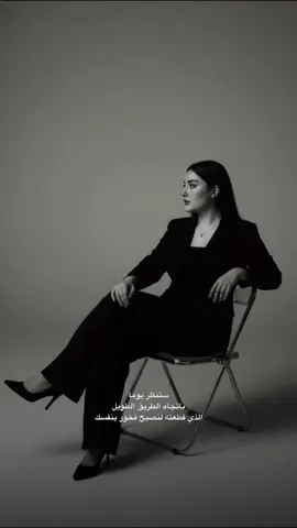 وصف عمل الصوره في gimini  Edit the photo into a black and white photo of the studio, showing a young woman sitting on a sleer in a folding metal chair, her legs crossed. She was wearing a black jacket, wide black cloth pants and black heels. Soft and simple lighting, producing a steady shadow and mood. Positioning the body relaxed, leaning slightly with one hand on the back of the chair, and the face inverted to the side. Simple background, simple and modern aesthetics 
