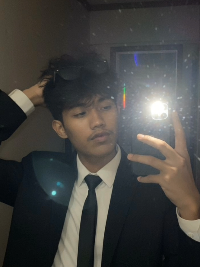 PROMPT: Subject: Person in the image [DO NOT CHANGE SUBJECT'S FACIAL FEATURES] A very ordinary iPhone mirror selfie with no clear subject or framing; a careless snapshot. The photo has a little motion blur and is slightly overexposed due to uneven light. The angle is awkward, the composition is non-existent, and the overall effect is aggressively mediocre - like an accidental photo taken while taking the phone out of your pocket to take a selfie. The subject takes a shaky iPhone mirror selfie at night, before he goes out. He wears a classic black suit jacket over a crisp white dress shirt, finished with a slim black tie that's slightly loosened at the collar and a sunglasses. The white shirt cuff peeks from the sleeve He is styling his hair. in a dimly lit room with no lights on except for the harsh phone flashlight, which creates stark highlights and blown-out reflections. The phone is very close to the mirror. The photo has a vertical aspect ratio of 9:16. Neon lights are faintly visible from the outside. #fyp #gemini #foryoupage 