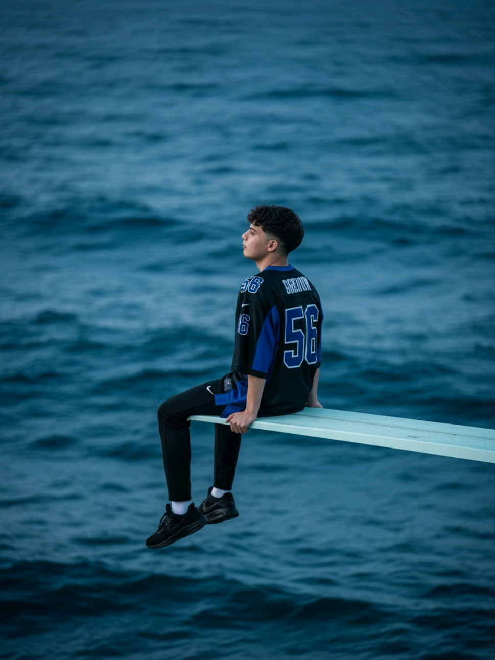 Crea tu propia fotografía con calidad de estudio usando Gemini AI.  Prompt en los comentarios.  Using this image as reference: generate a cinematic, editorial-style portrait of the same person, preserving facial structure, skin tone, hair texture, and emotional expression with maximum fidelity. The subject sits contemplatively at the far left edge of a thin white diving board that extends horizontally from the right side of the frame over a vast, textured deep blue ocean. The board resembles a minimalist surfboard—flat, clean, and perfectly horizontal, with no visible base or tilt. The ocean fills the entire background, with no horizon line, land, or sky visible—only endless waves in varying shades of blue and indigo. The subject is seated sideways, facing left, legs dangling straight down over the water. Both hands rest gently on the board, back straight, and head turned slightly to the right, gazing upward as if looking toward a sunset beyond the frame. The expression is calm, introspective, and emotionally resonant. The subject wears a black-and-blue sports jersey with the number “56” on the back, paired with black sport pants and black sport sneakers. The outfit is oversized and casual, evoking a sense of vulnerability and quiet strength. The hair and facial features must remain exactly as in the reference photo—no changes to shape, texture, or tone. Style: editorial portrait with cinematic, album-cover undertones.   Camera: full-frame DSLR simulation, 85mm lens, f/1.4 aperture, ISO 100, shutter speed 1/250s.   Angle: side-on view, camera positioned at water level or slightly above (5–10°), never from above.   Lighting: soft ambient light (~5200K), subtle reflections from the water, no direct sunlight.   Color grading: deep ocean blues, muted whites, soft contrast, slightly moody tones.   Resolution: 1080x1920, vertical format.   Mood: solitary, reflective, poetic. --v 5 --ar 9:16 --style raw --quality 2 --ref_photo #geminiai #prompt #inteligenciaartificial #photostudio 