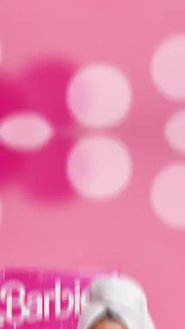 #Gemini Transform this selfie into a studio-quality portrait of an adorable 9yr old girl sitting on a hot pink small chair in a hot pink and white studio. The girl has sparkling eyes, and is joyfully smiling. She wears a premium white Bathrobe and a towel on her head. The background should feature soft pink lighting and glittery Barbie-themed props (mini purse, shoes, dress, makeup) and a life sized Barbie doll box (behind her) — hot pink with glitter, the white 'Barbie' logo at the top, and playful packaging details like stars, hearts, and shine. softly diffused by the bokeh. Use an 85mm lens aesthetic with very shallow depth of field, keeping the girl’s face and skin in sharp focus. Highlight hyper-realistic skin textures and The final image should be polished, cinematic, vibrant, and magazine cover–ready — a perfect blend of Barbiecore and luxury baby photography.