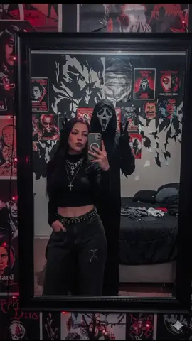 prompt: Gemini Crie uma foto de uma jovem mulher tirando uma selfie em frente ao espelho de um quarto com luz sombria e decoração alternativa. Ela usa um top preto curto e uma calça com espinhos, colares com cruz e maquiagem escura. Atrás dela, uma pessoa fantasiada de Ghostface, do filme Pânico, aparece de capuz preto, fazendo sinal de paz com os dedos. O quarto tem pôsteres de filmes ou arte sombria nas paredes, cama desarrumada e pequenas luzes decorativas, criando um clima retrô e levemente assustador.#gemini  #todomundoempanico 