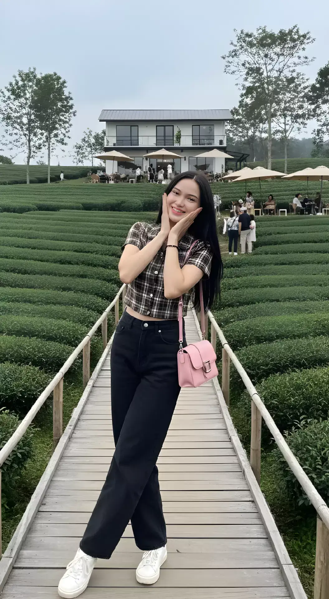 PROMPT : Foto realistis seorang wanita muda  dan memiliki rambut hitam panjang lurus. Ia memakai setelan: Atasan Model: Crop top dengan potongan pendek di atas pinggang. Motif: Kotak-kotak dengan kombinasi warna cokelat gelap dan garis tipis putih. Lengan pendek. Bagian depan ada kancing vertikal sebagai detail tambahan. Bawahan  Model: Celana jeans longgar. Warna: Hitam pekat dengan sedikit kesan washed. Alas Kaki Model: Sneakers kasual. Warna: Putih polos dengan sol tebal. Aksesori (Accessories) Tas: Shoulder bag. Model simpel dengan buckle silver. Gelang tipis berwarna hitam pada pergelangan tangan kiri. Tampilan Rambut & Gaya Umum Rambut: Terurai panjang, bergaya natural tanpa ikatan, sedikit berombak. Gaya: Casual chic dengan sentuhan streetwear minimalis. Latar Belakang Latar belakang menampilkan hamparan luas tanaman teh dengan pola barisan rapi yang membentuk lekukan mengikuti kontur tanah. Warna hijau pekat dari tanaman teh menciptakan suasana segar dan alami. Di tengah latar belakang, terdapat bangunan dua lantai bergaya modern-minimalis berwarna putih dengan atap hitam. Bagian bawah bangunan memiliki area outdoor café yang dipenuhi meja dan kursi, serta beberapa payung besar berwarna krem untuk tempat berteduh pengunjung. Terlihat juga beberapa orang sedang duduk dan berdiri di sekitar area café, menambah kesan hidup dan sosial pada suasana foto. Di sisi kiri dan kanan bagian depan, ada pagar kayu sederhana berwarna coklat alami yang membatasi jalan setapak dari papan kayu. Jalan kayu ini membentang lurus ke arah kamera, menjadi jalur utama yang menghubungkan café dengan perkebunan teh di sekitarnya. Bagian langit di belakang tampak berawan tipis, dengan pohon-pohon tinggi menjulang di area belakang café, memberi nuansa alam pedesaan yang asri. Latar ini secara keseluruhan menghadirkan suasana alami, segar, dan tenang, dengan perpaduan antara hamparan hijau perkebunan teh dan bangunan modern café yang menjadi daya tarik utama wisata. Deskripsi Pose Berdiri di atas jembatan kayu kecil di tengah perkebunan teh. Tubuhnya sedikit condong ke satu sisi, dengan berat badan lebih bertumpu pada kaki kanan, sementara kaki kiri diletakkan agak ke depan dengan posisi silang ringan sehingga memberi kesan santai dan feminim. Kedua tangannya diangkat ke area wajah, dengan kedua telapak menempel pada sisi pipi kanan dan kiri, seperti gaya ekspresi tersenyum malu atau gemas. Wajahnya menghadap lurus ke arah kamera dengan ekspresi tersenyum ceria. Posisi bahu tampak sedikit rileks, sementara kepalanya tegak dengan sedikit condong ke arah kanan, menambah kesan natural. Aksesori berupa tas kecil berwarna pink digantung di lengan kiri, dibiarkan jatuh ke samping paha, mengikuti postur santainya. #gemini #ai #cafe #trending #outfit 