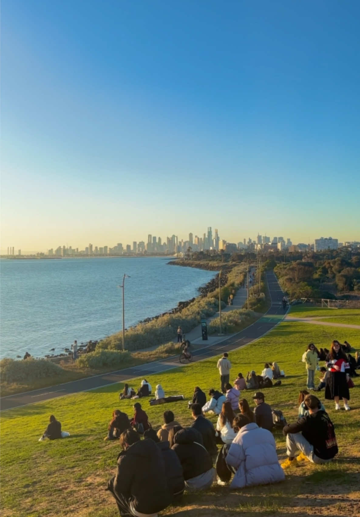 What's your perfect kind of afternoon? Ours can look a little like this... 🙌 🎥: @Love, vina ✨🦋  📍: Point Ormond Lookout, Narrm (@Visit Melbourne) #SeeAustralia #ComeAndSayGday #Melbourne #Travel #TravelTok ID: A group of people sitting on a grassy hill overlooking the ocean and the Melbourne city skyline at sunset.
