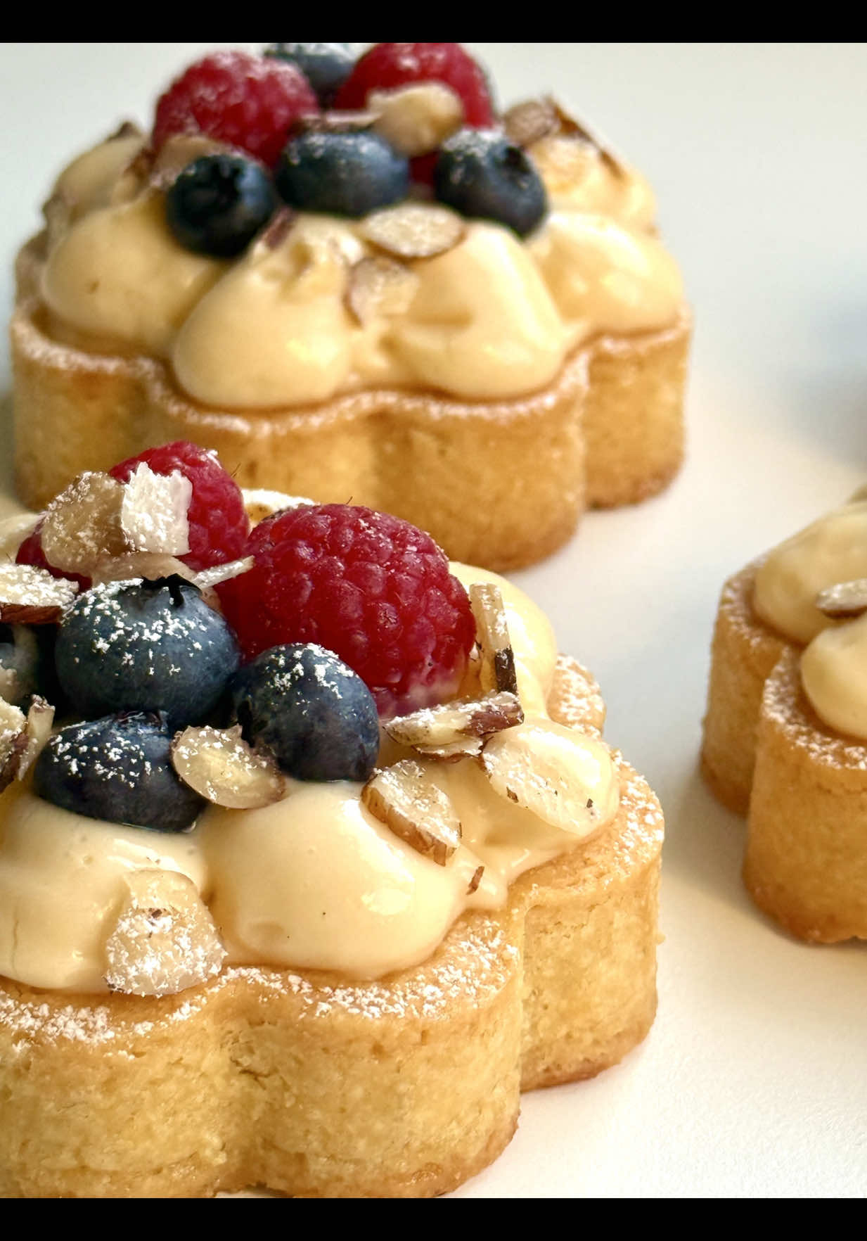 Ad | Berry Tartlets Recipe 🫐🤍 These tartelettes are delicate little treats in a floral shape . They look as if they came straight from a French patisserie.🥰 📝 Ingredients (for 4 tartelettes, Ø 9–10 cm) For the shortcrust pastry: 	 •	150 g all-purpose flour (1 ¼ cups) •	75 g cold butter, cubed (5 ½ tbsp) •	50 g sugar (¼ cup) •	1 egg yolk •	1 tbsp cold water •	1 pinch of salt For the filling: 	 •	4–5 tbsp fruit jam (e.g. homemade plum jam or any jam of your choice) For the pastry cream: 	 •	250 ml milk (1 cup) •	2 egg yolks •	40 g sugar (3 tbsp) •	20 g cornstarch (2 ½ tbsp) •	1 tsp vanilla extract •	30 ml heavy cream, liquid (2 tbsp) For decorating: 	 •	about 100 g fresh blueberries •	about 100 g fresh raspberries •	a few hazelnut slices •	powdered sugar for dusting 🤍 Instructions 	 1.	Mix flour, sugar and salt. Add cold butter and rub into crumbs with your fingertips or a mixer. Add egg yolk and water, then quickly knead into a dough. Wrap in cling film and chill for 30 minutes. 	 2.	Preheat the oven to 180 °C / 350 °F (top/bottom heat). Grease the molds. Roll out the dough and cut circles for the bases. Cut strips for the sides, measure them and place into the molds, pressing them gently with your fingers (not too firmly). Prick the base with a fork, line with baking paper and fill with baking beans. Blind-bake for 15 minutes. Remove the beans and bake for another 15 minutes until golden brown. Let cool completely and carefully remove from the molds. 3.	Heat the milk with vanilla extract. In a bowl, whisk egg yolks, sugar and cornstarch. Slowly pour in the hot milk while stirring, then return everything to the pot and cook, whisking constantly, until thickened. Transfer to a bowl, cover the surface with cling film and let cool completely. Once cooled, add the 30 ml liquid cream and beat with a hand mixer until smooth and creamy. 	 4.	Spread jam over the cooled tart bases. Spoon pastry cream on top and smooth it out. Pipe lines from each petal edge toward the center, creating a flower pattern. 	 5.	Decorate with blueberries, raspberries and hazelnut slices. Dust with powdered sugar just before serving. A charming, fruity and creamy delight 🤍