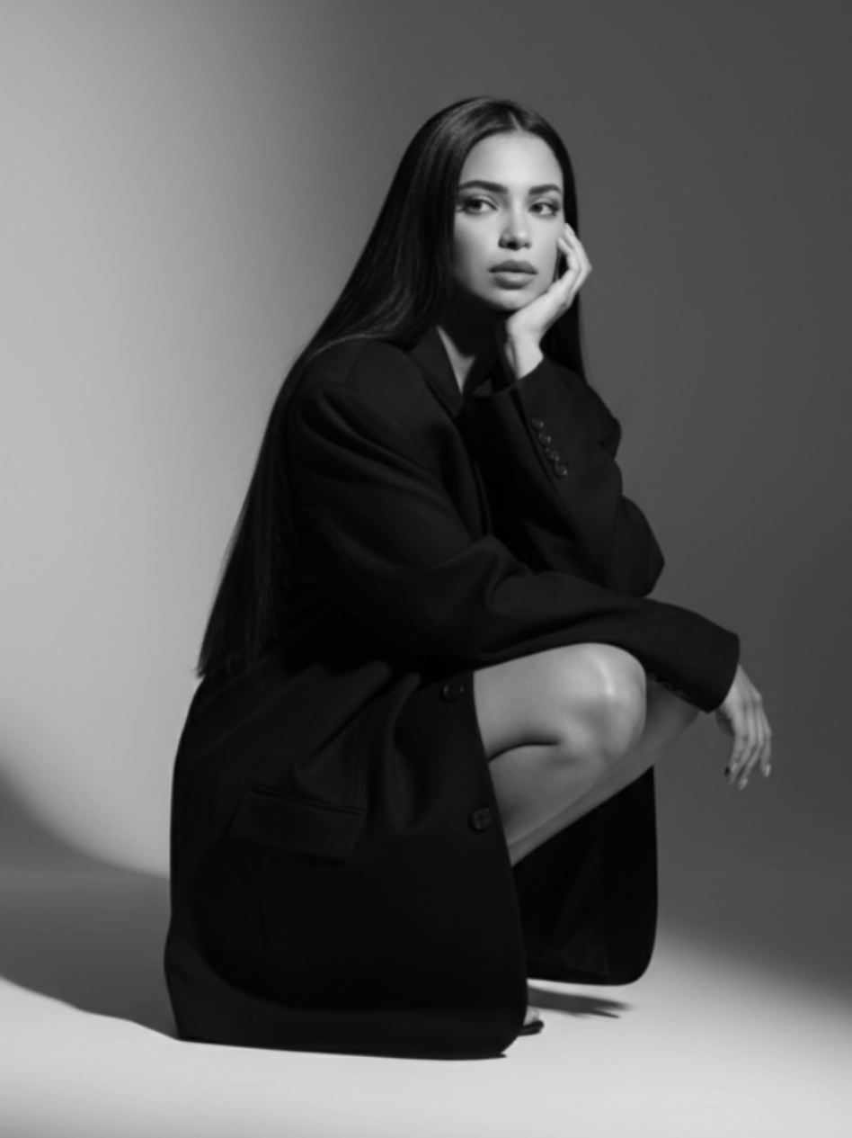 Texto do Gemini para foto profissional Create an artistic black and white portrait of this woman with a pensive expression, posing in a crouched position. She wears an oversized black blazer that covers part of her legs. Her hair is long, loose, and flowing, falling over her shoulders. Her right hand gently supports her face, while her gaze is turned to the side, conveying introspection and specificity. The lighting is soft, casting striking shadows on the wall behind her, creating a minimalist and sophisticated atmosphere #ViralCut #IA #gemini #foryou #passoapasso 