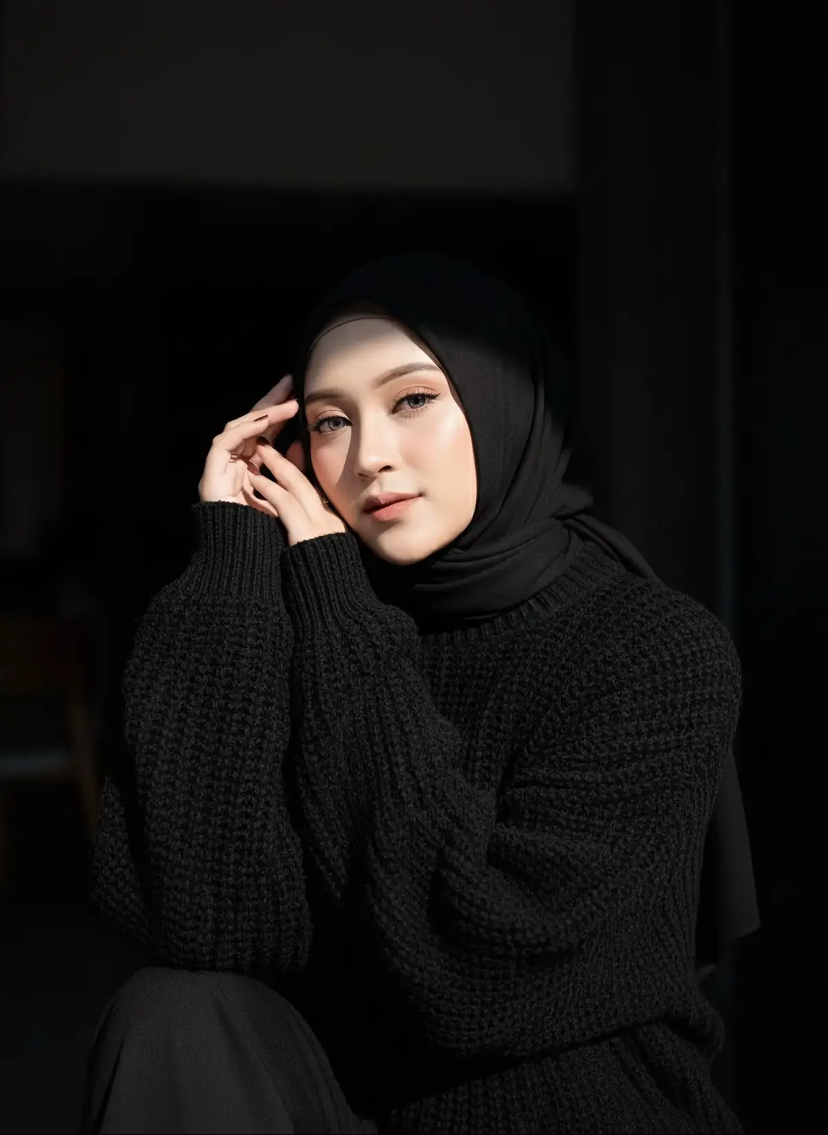 Prompt :  Don't change her face, Artistic fashion portrait of a young woman in a luxurious chunky knit black sweater with voluminous textured yarn. She is framed in dramatic side lighting, a narrow beam of sunlight cutting across her face to create a striking chiaroscuro effect. The background is deep and dark, almost black, emphasizing the play of light and shadow. Her expression is serene and introspective, with delicate natural makeup and softly. ~~The overall atmosphere is minimalistic, moody, and elegant, reminiscent of high-fashion editorial photography for a luxury magazine. She's wearing pashmina hijab black, she's wearing sweater fleece aesthetic black with ribbed in hands Sitting or semi-reclining, shoulders slightly tilted to create dimension. • Hands: One arm is raised above the head with the fingers forming a graceful curve; the other hand gently supports the chin or cheek. • Head: Slightly tilted to the side, eyes directed toward the camera with a calm, soft gaze. • Expression: Neutral, slightly melancholic or contemplative. — Mood & Lighting • Lighting: Single-source natural light from a narrow window or a no in the curtains, creating a thin line ht that cuts across. #ai #geminiai #aitrend #aiart 