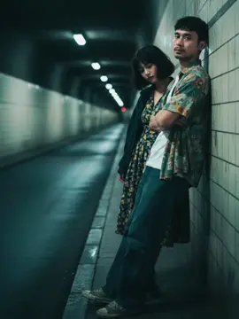 prompt: Cinematic night scene in a dimly lit tunnel. Two young people (use reference photo for accurate face) stand against the tiled wall. A man with a messy french crop-hair, wearing a patterned button-up shirt and loose blue jeans, leans casually against the wall with arms crossed, gazing forward with a calm, confident expression. Beside him, a young woman with short wavy hair in a dark jacket and floral dress leans forward slightly towards the road, head tilted,expression soft and tired yet intimate, eyes half-closed. The tunnel is lined with glowing overhead lights stretching into the distance, perspective drawing deep into the vanishing point. Wet asphalt reflects faint highlights. The atmosphere is moody, nostalgic, and cinematic.Color tones muted teal, green, yellow and amber, with subtle film grain and soft vignette. Camera angle mid-shot, slightly low, capturing both figures with the tunnel receding behind them. Wong Kar-wai style, urban melancholy, intimate yet detached. #lewatberanda #gemini #4u 