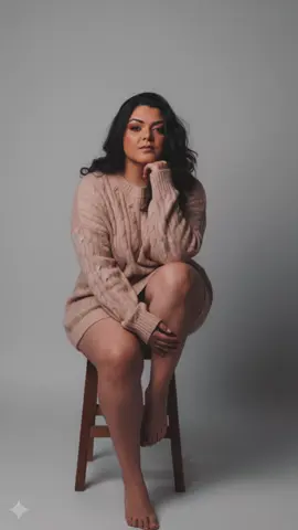 Crie um retrato de filme hiper-realista e dramático. Um retrato vertical (8K) da pessoa na foto de referência, certificando-se de que todas as características faciais combinam perfeitamente com a foto original. Ajuste os detalhes da pele facial e das pupilas dos olhos. Uma mulher com cabelos castanhos escuros, está sentada em um pequeno banquinho. Ela está vestindo um suéter bege claro folgado. Sua expressão facial é calma e atraente e sonhadora, com o olhar fixo na frente. Sua mão direita segura o queixo e a esquerda segura o tornozelo. Ela está sentada com as pernas cruzadas sobre o ombro. Seu corpo é bem proporcionado, com pernas longas, brancas e finas. O fundo é um cinza claro suave, criando um contraste sutil com a cor do suéter. A iluminação é suave e ligeiramente escura, criando uma atmosfera tranquila e introspectiva. A perspectiva é ligeiramente superior, proporcionando uma perspectiva profunda. A composição da imagem é centrada no assunto, com foco principal nas pernas finas, expressão e pose da mulher As cores suaves e o foco na textura do suéter de malha adicionam calor e beleza visual. Este estilo de fotografia é artístico e elegante, com ênfase nos detalhes sutis e na beleza natural. Mostra uma imagem de todo o corpo feminino, da cabeça aos pés. Mantenha o corpo plus size, seios grandes, e não mude o rosto da foto enviada #iaplus #gemini #ensaiofotografico #fotoia 