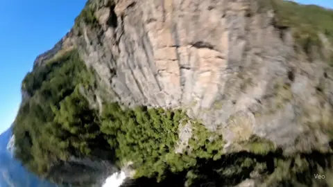 Ultra realistic cinematic FPV POV of a wingsuit dive along a steep mountain ridge. Aggressive nosedive down a sheer cliff face at insane speed, hugging the rock surface. Sharp right bank skimming treetops, then a hard left roll threading through narrow canyon walls. Low sweep across a waterfall mist, droplets hitting the lens POV. Final steep climb into the open sky with a wide mountain vista. Pure camera flight — no drone body, no propellers, no gimbal, no shadows, no reflections. #ai #aicreator #drone #fvp #fyp 
