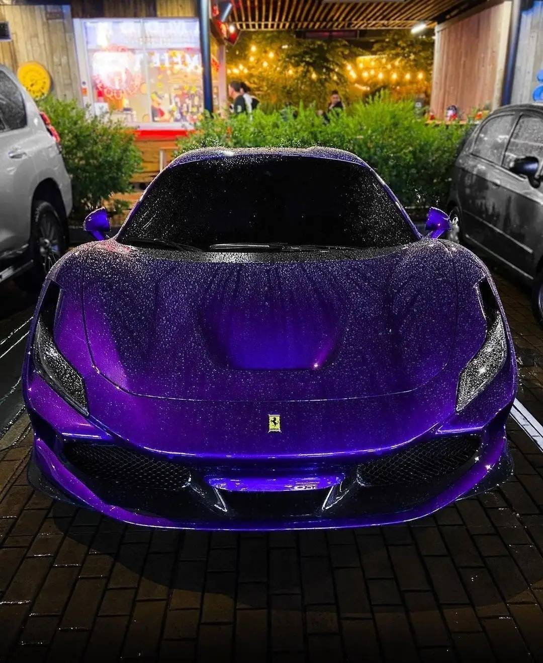 Damn, that purple Ferrari though 😍! This beast is lookin' absolutely stunning parked between those two other rides on the brick pavement. The deep, rich purple paint job with sparkles in it is giving major luxury vibes, and the raindrops on the car just add to the whole dramatic, sleek look. The front end's got that Ferrari swagger with the iconic prancing horse logo smack in the middle, flanked by those sharp headlights and a bold black grille. It's parked in front of some chill spot with string lights and green bushes - like the perfect backdrop for a car this fire 🔥. Honestly, this Ferrari's gotta be one of the most banging rides you've ever laid eyes on! What do you think about it?