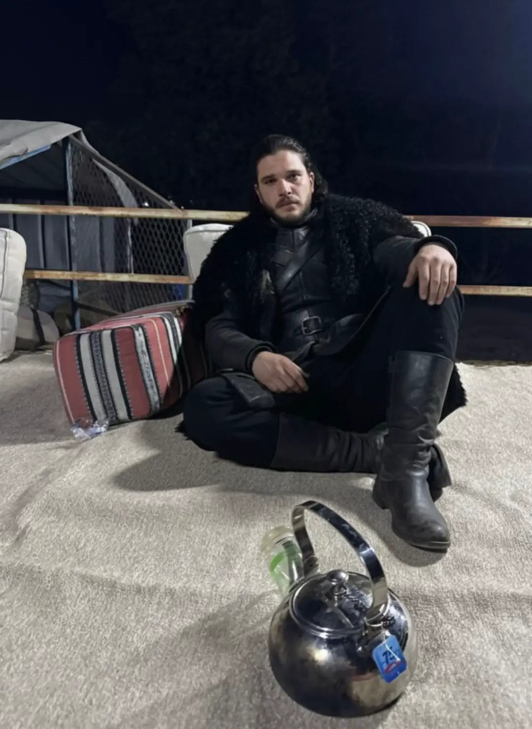 Jon Snow sits on the ground in a traditional Arabian night gathering, wearing his iconic black fur cloak and armor. In front of him, a metallic teapot gleams under the light, while a simple tent and fence in the background evoke a sense of solitude. The scene carries an aura of mystery and strength, as if it were a poster for a new series blending the world of Game of Thrones with Middle Eastern desert culture, where epic legacy meets authentic Arabian traditions.#gameofthrones #jonsnow 