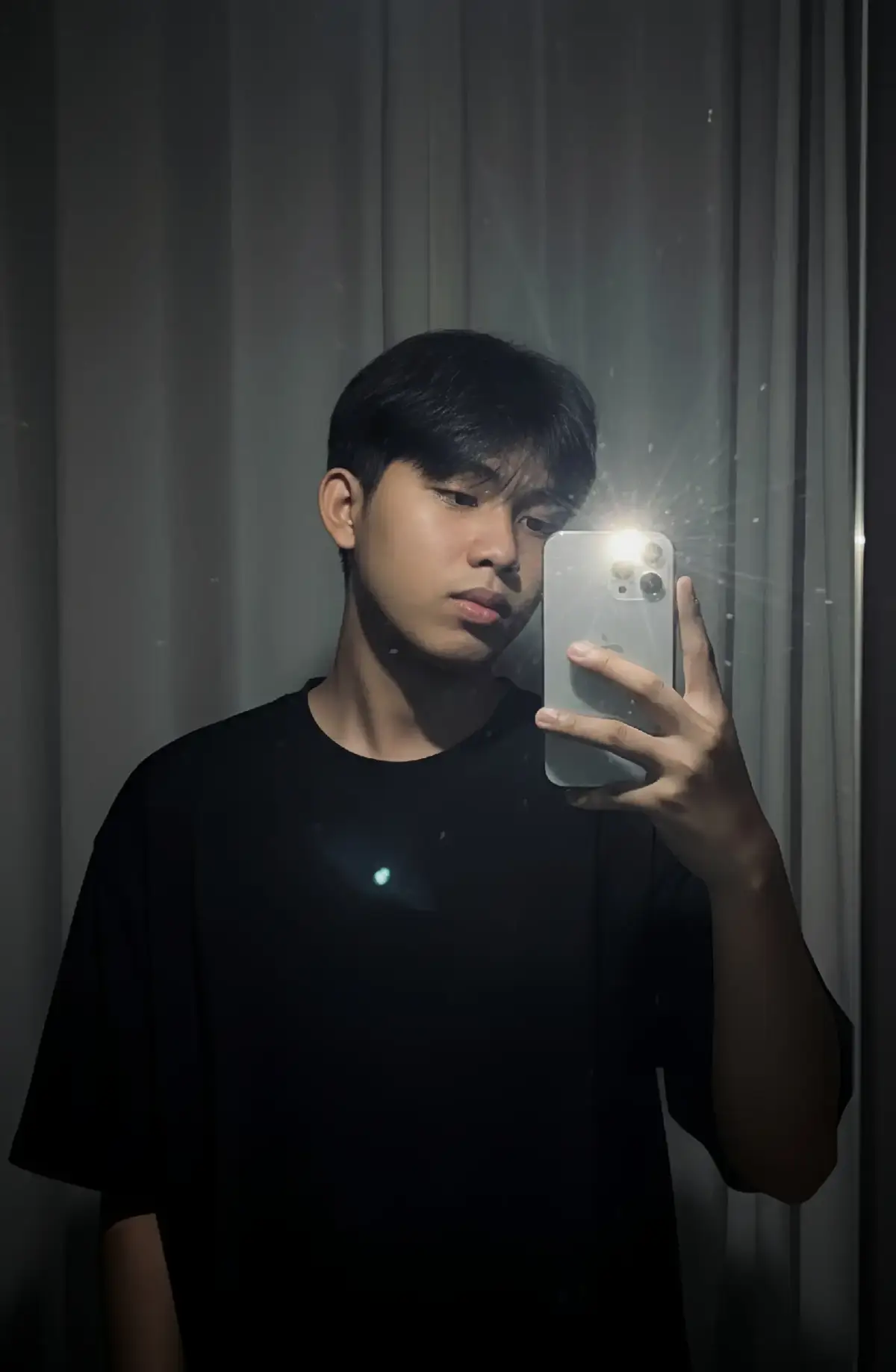 Buatlah foto ini sedang berfoto mirror di kaca dengan menggunakan hp iPhone 11 Pro Max dengan flash, sehingga menghasilkan foto yang aesthetic. UNTUK FOTO UTAMA: Buatlah gambar yang diambil seperti foto biasa, tanpa subjek atau properti yang jelas. Foto tersebut harus memiliki sumber cahaya yang konsisten, seperti lampu kilat dan ruangan gelap, yang tersebar di seluruh foto. Jangan rubah wajah dan rambut orang tersebut. Ganti latar belakang di belakang orang tersebut dengan tirai warna putih. Gunakan kaos hitam oversize untuk memberi kesan kasual dan modern #gemini #ai #prompt #cgpt #fyyypppppppp