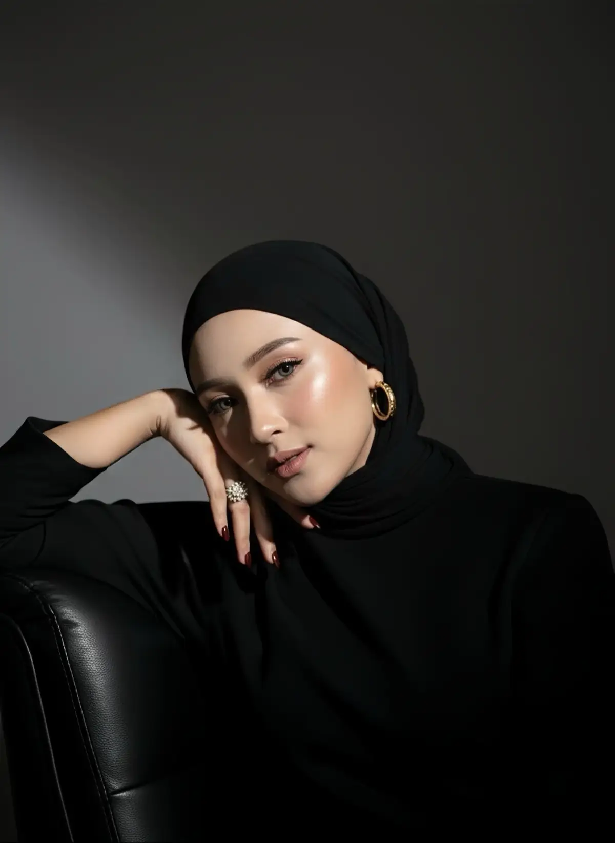 Prompt : Create don't change the face. Quality HD. Close-Up portrait of a woman wearing a black hijab and a black top, seated and resting her head on her hand. She has noticeable, glowing makeup with a highlight on her cheekbone and is wearing gold hoop earrings and a large, ornate ring on her finger. The lighting is dramatic, with a single, strong light source illuminating her from one side, creating high contrast against the dark gray, plain background. The overall mood is elegant and intense. #ai #aitrend #aiart #gemini #geminiai 