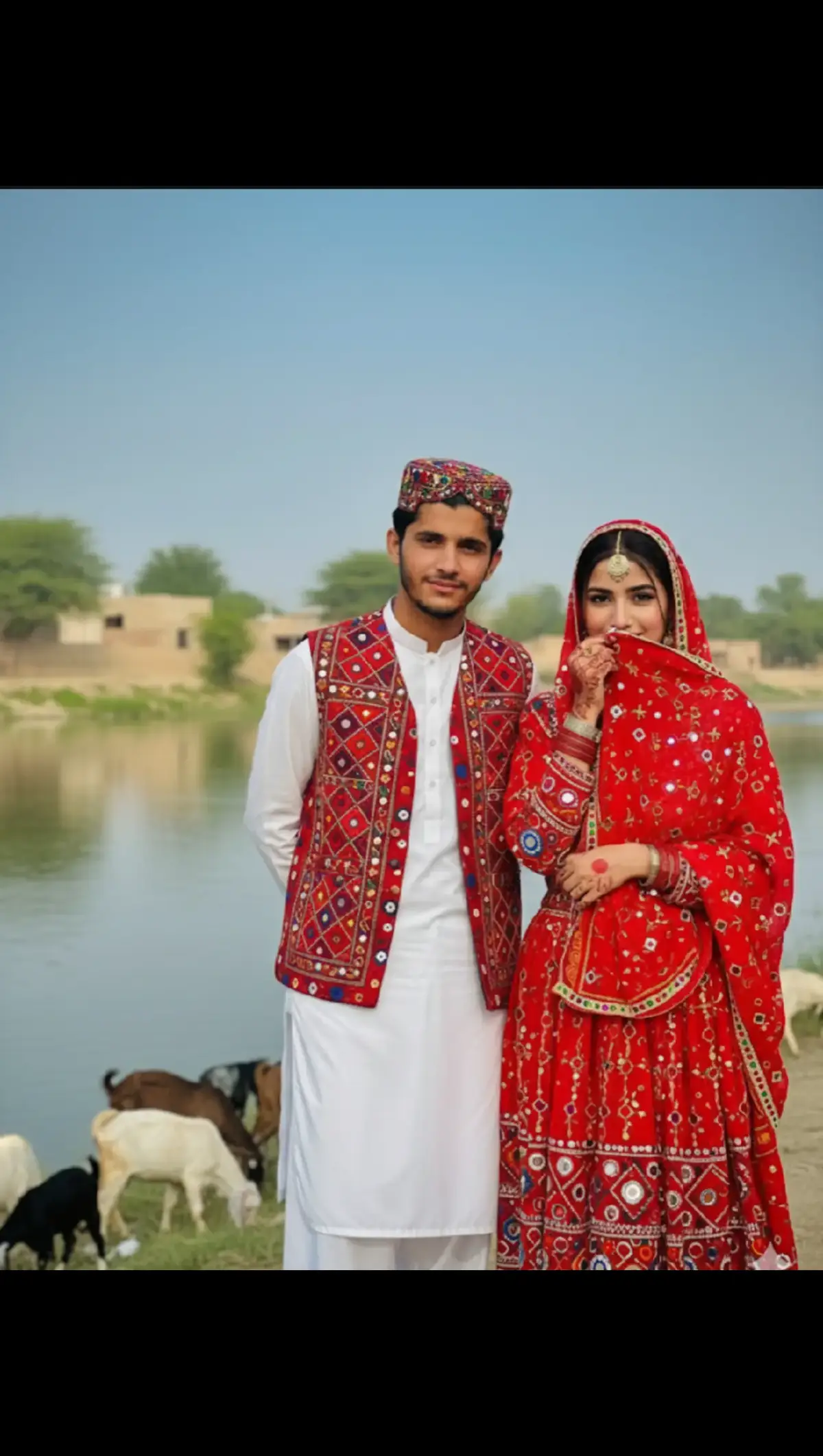 realistic and cinematic portrait based on uploaded selfies (strictly keep original faces with no changes, preserve real skin tone and natural colors). Scene captured at the Sindhi river bank with goats around, full of cultural vibes. The male (me) is wearing a pure white Sindhi traditional shalwar kameez, with Sindhi topi and Ajrak patterned waistcoat, slightly smiling in a natural way. My friend (based on uploaded actress selfie) is in 1990s Sindhi dress with handmade art designs of sheshay and motiyan, holding her dupatta with teeth to shyly cover part of her face, giving a shy-smiling expression. Both look natural, emotional, and connected in the cultural environment. Background shows peaceful river, goats, and village trees, with soft natural light. Ultra realistic, hyper detailed textures, traditional Sindhi culture aesthetics, sharp focus, perfect color grading. Add watermark text 