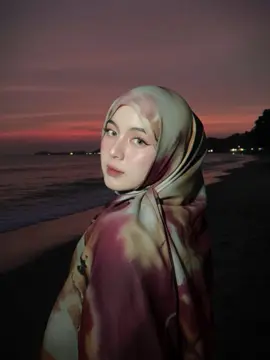 maintaining the exact same face and feature as the reference photo, close up photo, make me look im on a beach where it's dark but someone took my picture with a flash and my face is a bit glowing. in the background add the sunset like the sun has 98% set leaving purple red sky and it's almost dark the image is taken from behind my back,the eyes looking at camera,and I've been told to picture for the picture im wearing a loose flowy selempang belakang style shawl 1/2 #geminiai #sunset #pantai 