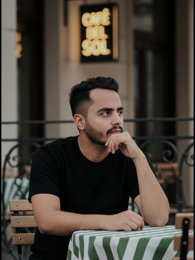 #chatgpt #GeminiAI #Prompt 25 year old young man with a stylish haircut, wearing a black pant with t-shirt  embroidered details, sitting at an outdoor cafe table. He is leaning his chin on his hand, looking thoughtfully to the right. The table has a green and white striped tablecloth, and there are blurred green leaves in the foreground, creating a natural frame. In the background, other cafe chairs and a dimly lit sign are visible. The lighting is soft, suggesting a pleasant outdoor setting. 100% matching my face #trending #foryou 