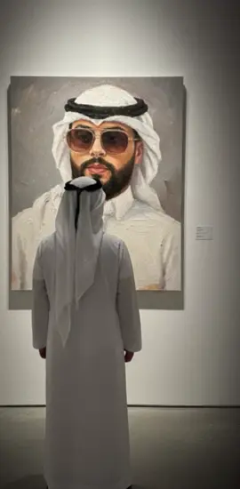 الطريقة تحمل التطبيق Gemini.  بعدين تحط  Using the attached image as a reference for the man’s face and appearance, create a scene in which the man stands in a modern art gallery, seen from behind, and gazes at a large oil portrait of himself on the wall. The painting should depict the same man in a realistic and expressive oil painting style, using textured brushstrokes and soft colors on his face and upper body. The gallery wall is clean and white, and a soft spotlight illuminates the artwork, creating a professional exhibition atmosphere. Keep my original look exactly the same.
