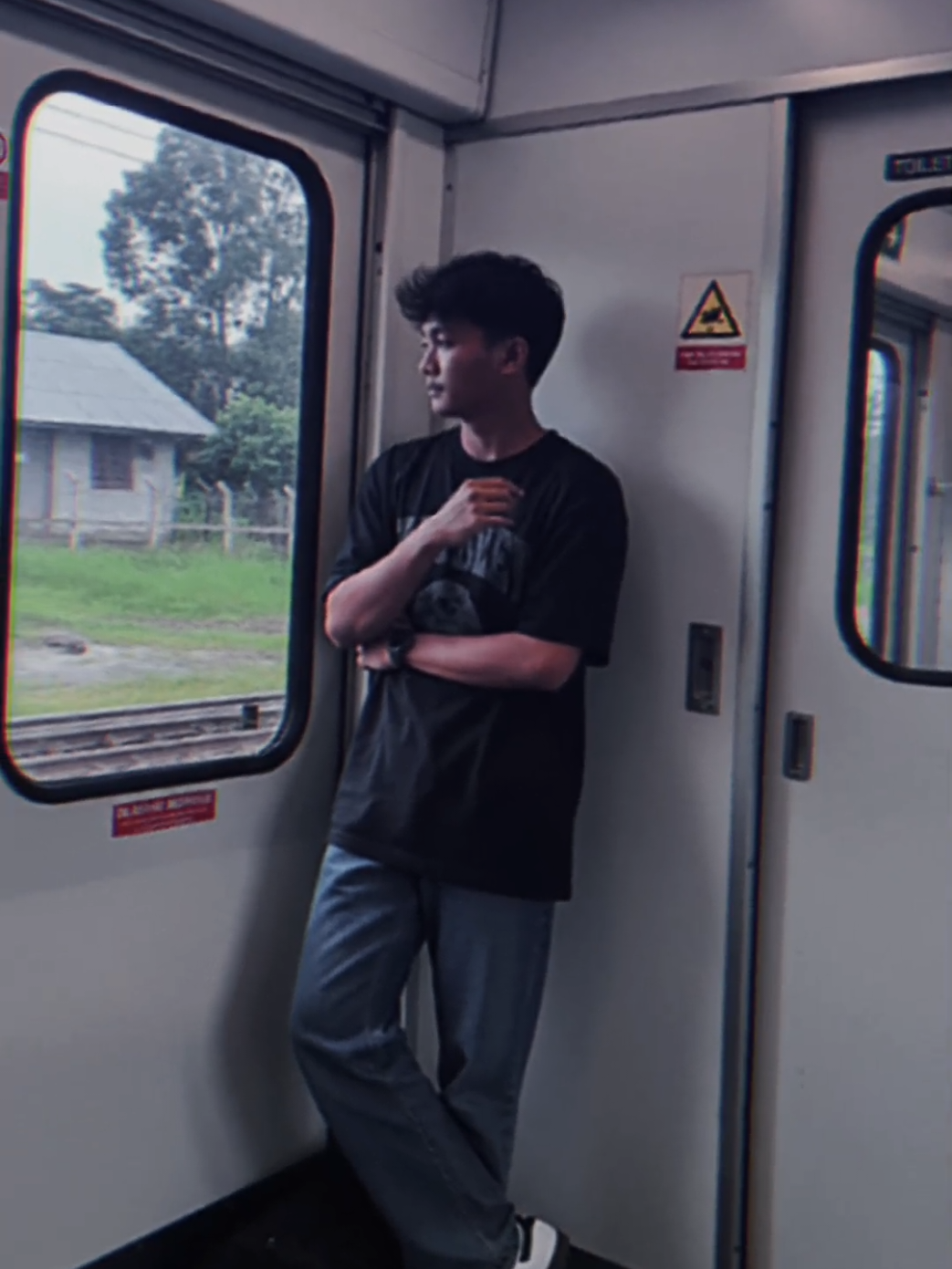 Ubah foto yang saya unggah tanpa mengubah wajah dan gaya rambut sedang berdiri di dalam kereta, mengenakan kaos oversize berukuran XL dengan sablon abu abu .celana panjang jeans gombrang dan sepatu Adidas samba klasik warna putih Di bawah nya terdapat barell bag Posisi tubuhnya bersandar pada dinding pintu kereta, tangan kanan terlipat ke dalam, tangan kiri melipat ke luar. Kepalanya lurus sedikit menunduk ke arah kiri, pandangan diarahkan keluar jendela, dengan rambut sedikit berantakan terkena angin.  Pose: Berdiri menyilang santai, punggung bersandar pada pintu, kaki kiri sedikit maju ke depan, kedua tangan menyilang. Sisi kanan gambar: Pintu toilet dengan tulisan “TOILET 1”, tanda darurat berwarna kuning segitiga dengan ikon bahaya, serta kotak kecil merah bertuliskan “TARIK BILA ADA KEBARAKAN / PULL IN CASE OF FIRE”. Ada juga sebagian pintu yang terbuka ke arah lorong kereta. Sisi kiri gambar: Pintu darurat kereta dengan jendela besar persegi panjang, terdapat stiker “DILARANG MEROKOK” dan tulisan merah kecil lain di bawahnya. Melalui jendela, terlihat suasana luar berupa rumput hijau, rel besi yang ditumpuk, pagar, pepohonan, dan sebuah bangunan atap seng abu-abu. Latar belakang: Interior kereta dengan dominasi warna putih dan abu-abu, pencahayaan dari lampu neon di atas, serta pemandangan luar jendela yang terlihat jelas berupa area hijau dengan rel dan pepohonan basah seperti setelah hujan. Gambar di ambil menggunakan kamera iphone dengan kualitas resolusi agak rendah, kontras rendah, bayangan lembut, tidak ada efek estetika tambahan, foto terlihat original tidak seperti di edit. Rasio 3:4 potrait #ai #gemini #promt 