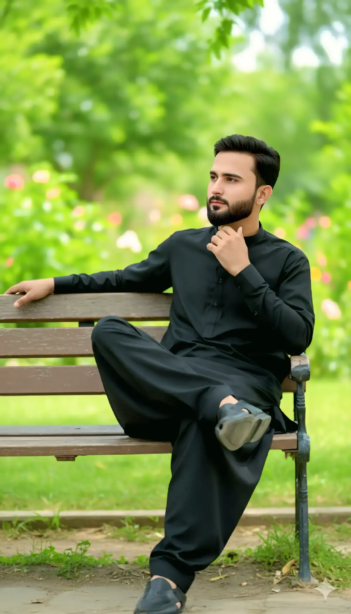 Full-body ultra-realistic portrait of the same young man (use reference face and hairstyle). A handsome South Asian man in his late 20s to early 30s, with short, dark hair and a well-groomed beard, wearing a traditional black shalwar kameez.  He is seated on a rustic wooden bench, leaning back slightly with his left arm resting on the backrest, and his right hand gently touching his neck/jawline. His legs are crossed at the ankles, with his left foot slightly elevated. He has a relaxed yet thoughtful expression, looking off to the side (to the left of the frame).  The background is a soft-focus, vibrant green garden with lush foliage, suggesting a sunny outdoor setting. The lighting is natural and bright. Realistic, cinematic photography.