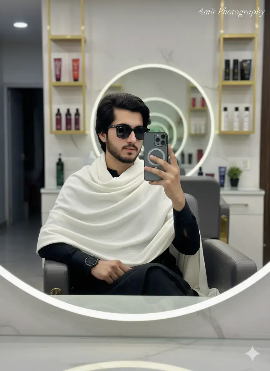 Ultra-realistick cinematic photo of a stylish young boy sitting on a luxury salon chair in front of a big mirror, holding an iPhone 17 pro max and clicking a selfie. He is wearing a same dress white shawl draped over his shoulders. black sunglass watch, giving a confident fashionable vibe The baci shows salon interiors with glowing vanity lights, hair products on the counter, and glossy reflection in the mirror. the scene has a cinematic lifestyle tone, shot with a sony A7R IV, 35mm f/ 1.4 lens. Note: use facial description from the uploaded image.