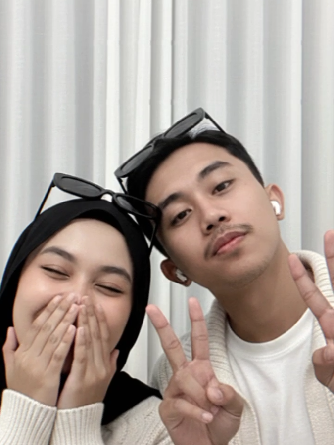 Edit foto saya tanpa merubah wajah  Selfie close-up pasangan muda di dalam ruangan. Di sisi kiri, seorang wanita muda berhijab hitam, yang di atas kepalanya disematkan kacamata hitam stylish. Dia mengenakan jaket atau sweter rajut berwarna putih tulang dengan tekstur yang terlihat jelas. Ekspresi wajahnya terlihat serius namun tenang, dengan pandangan mata langsung ke arah kamera. Dia menyentuh pipinya dengan tangan kiri, menambah kesan kasual. Di sisi kanan, seorang pria muda  kacamata hitam bertengger di atas kepalanya. Dia memakai kaus putih di bawah jaket atau sweter rajut berwarna putih tulang yang senada dengan pasangannya. Pria ini berpose dengan jari telunjuknya menunjuk ke arah kamera (atau membentuk gestur 